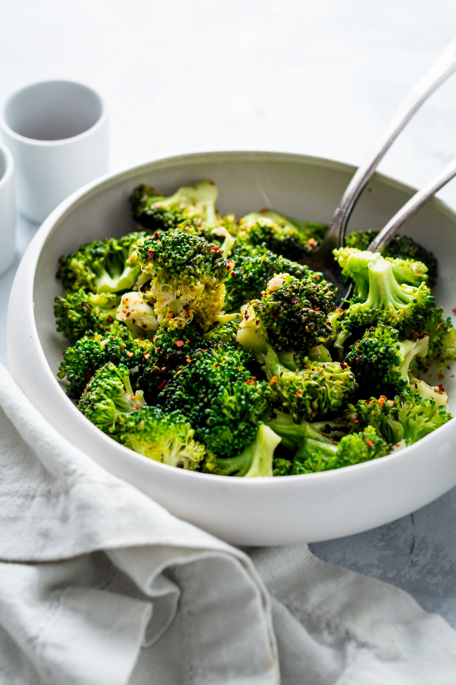 Crispy pan fried broccoli in a white bowl with a white napkin and two serving spoons.