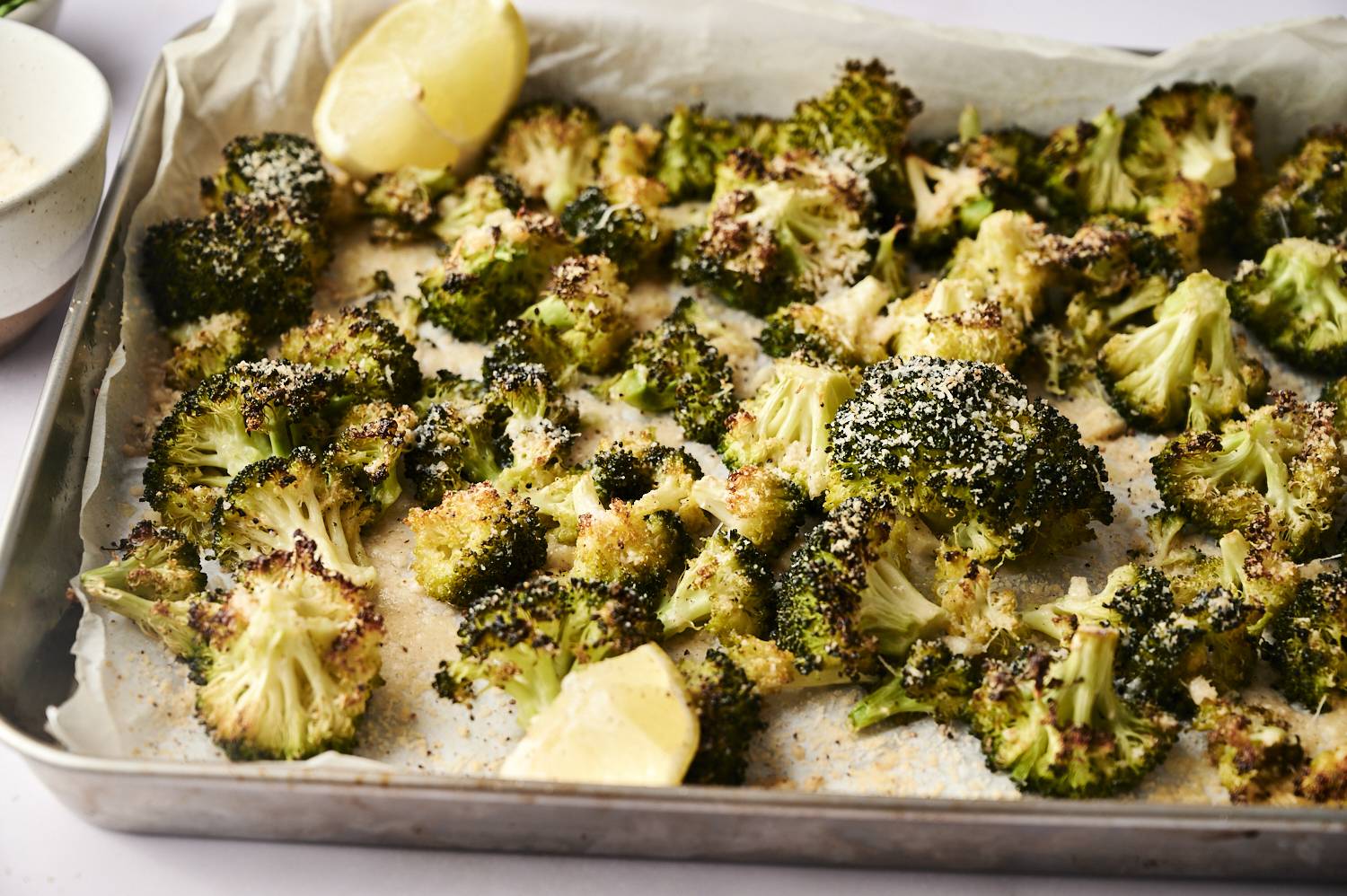 Roasted broccoli with crispy edges and parmesan, garnished with lemon wedges on a baking sheet lined with parchment paper.