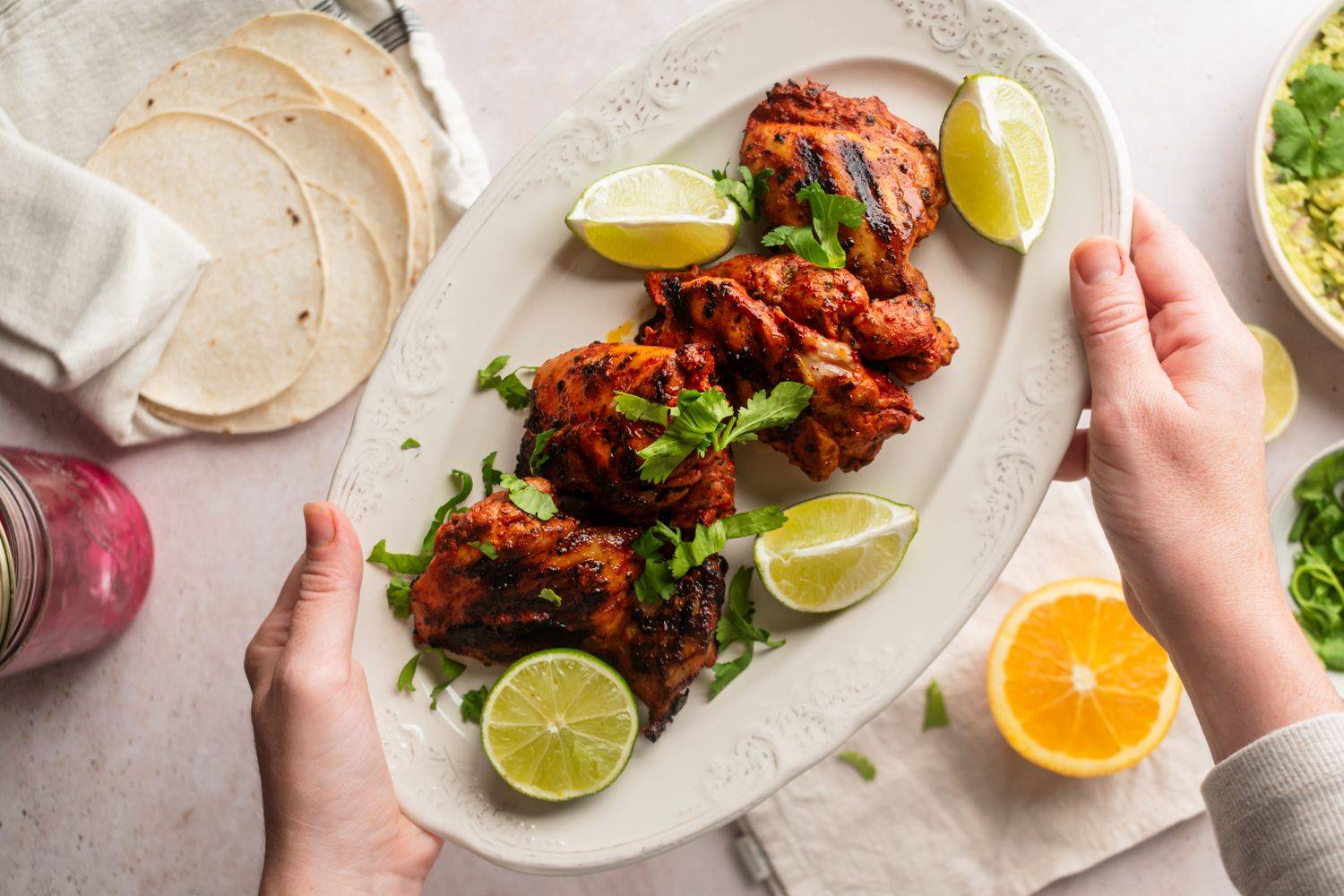 A hand holds a platter of grilled chicken garnished with lime and cilantro, alongside tortillas and fresh ingredients.