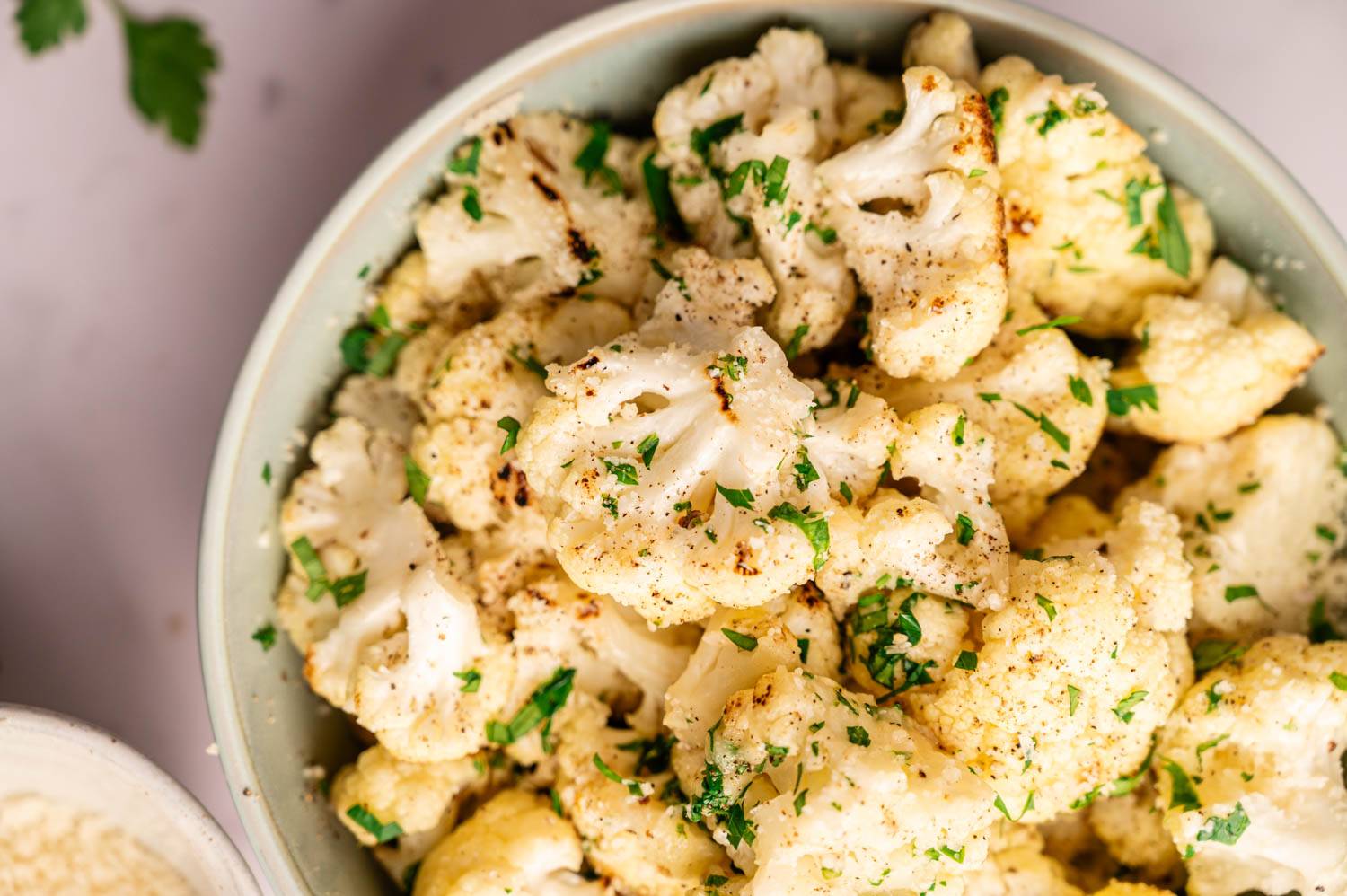 A bowl of roasted cauliflower florets garnished with fresh parsley sits on a table. The cauliflower is lightly browned, seasoned with pepper.