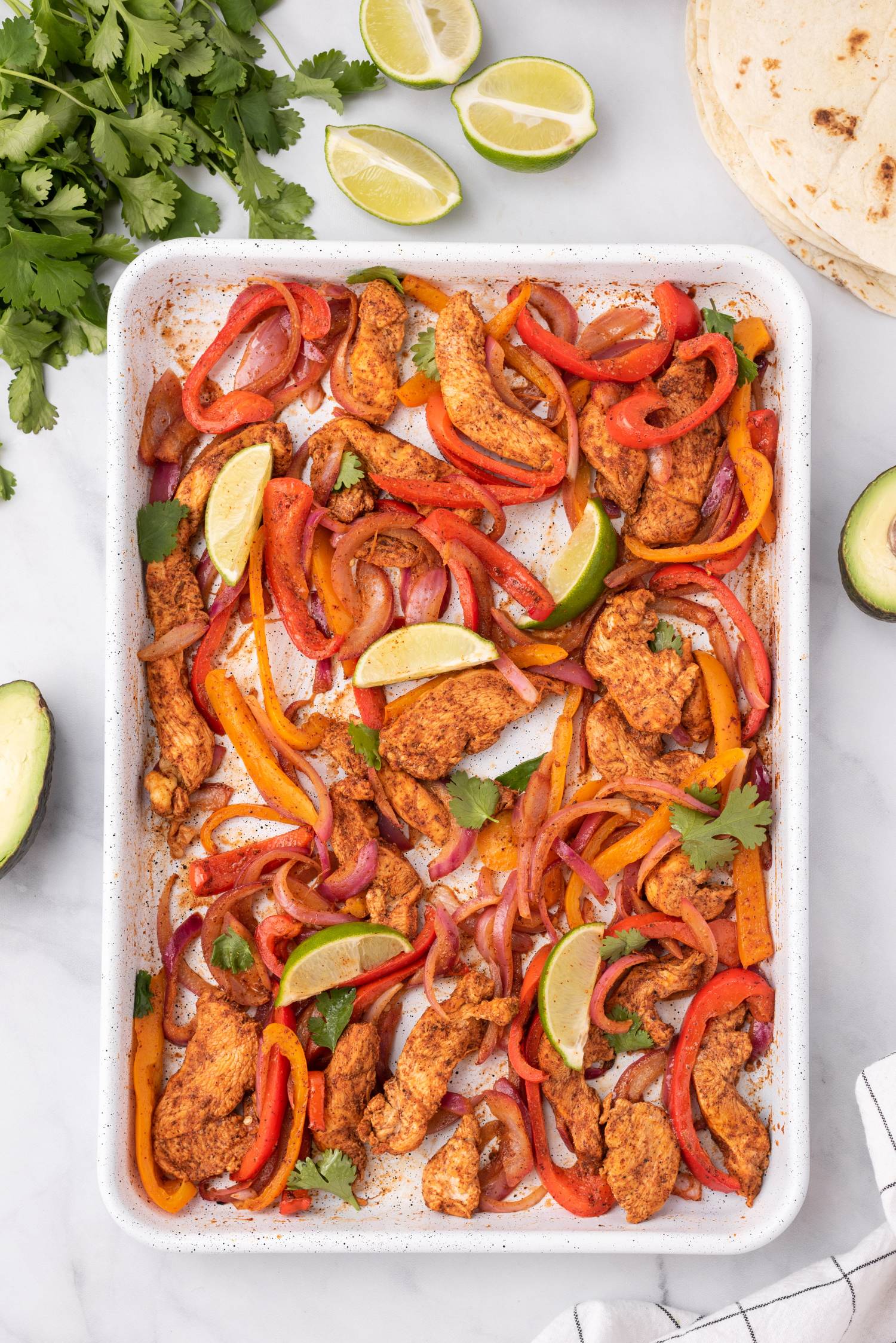 Sheet pan with fajita chicken, bell peppers, red onions, lime wedges, and fresh cilantro.