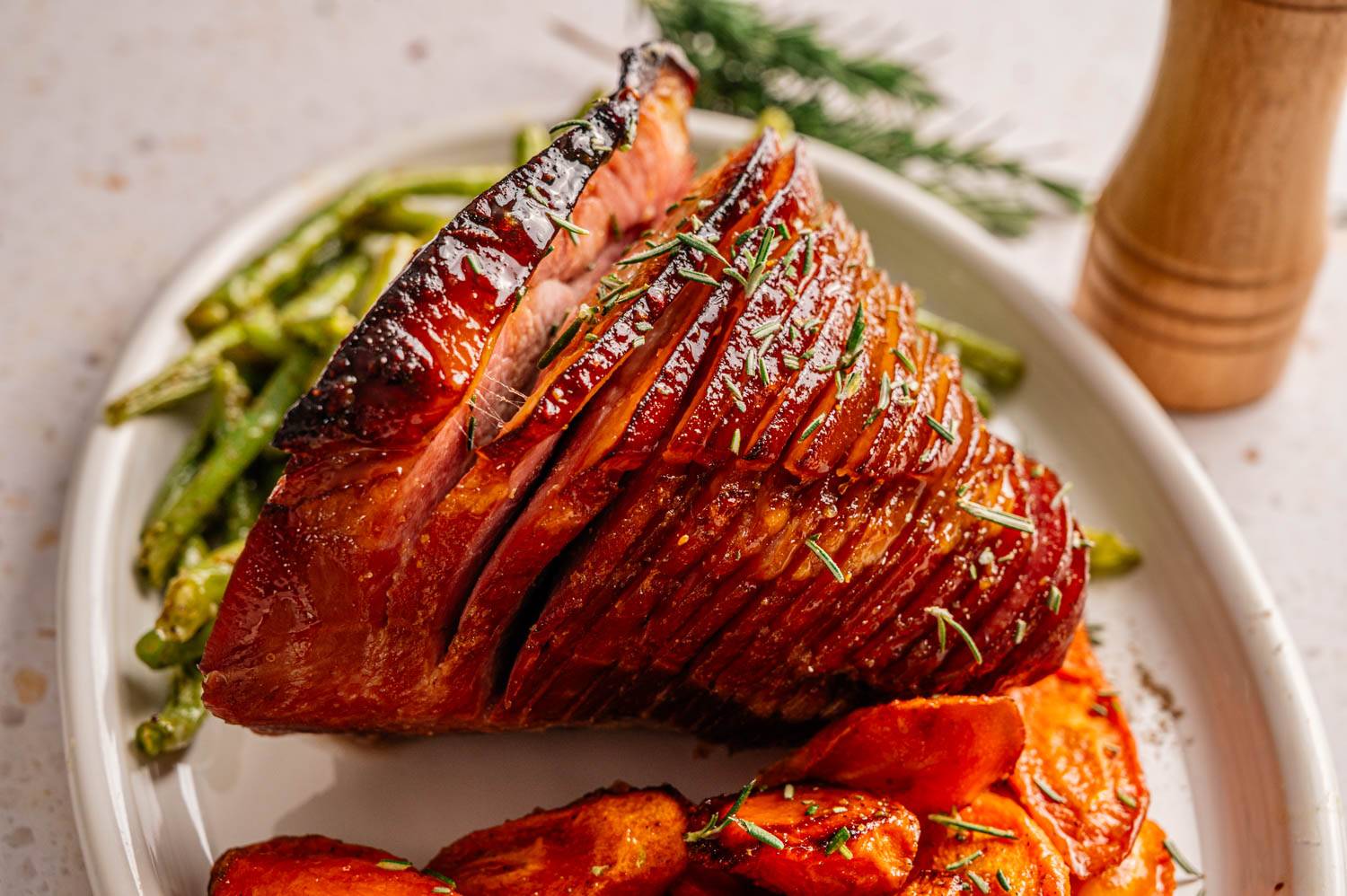 Sliced glazed ham garnished with rosemary on a white plate, accompanied by roasted carrots and green beans. A pepper mill adds warmth to the setting.