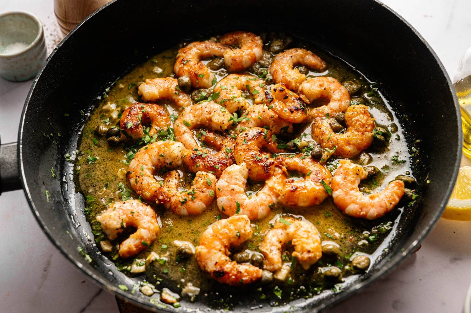 Pan-seared shrimp in a garlic and herb sauce in a black skillet. Golden brown crust, garnished with capers and parsley, creating a savory, appetizing scene.