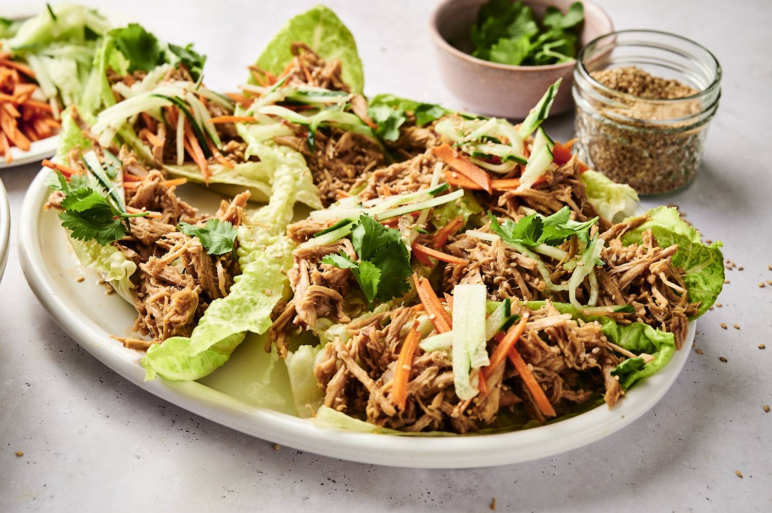 Lettuce wraps filled with shredded slow cooker Banh Mi pork, topped with matchstick carrots, cucumber strips, fresh cilantro, and sesame seeds, arranged on a white platter with jars of toppings in the background.