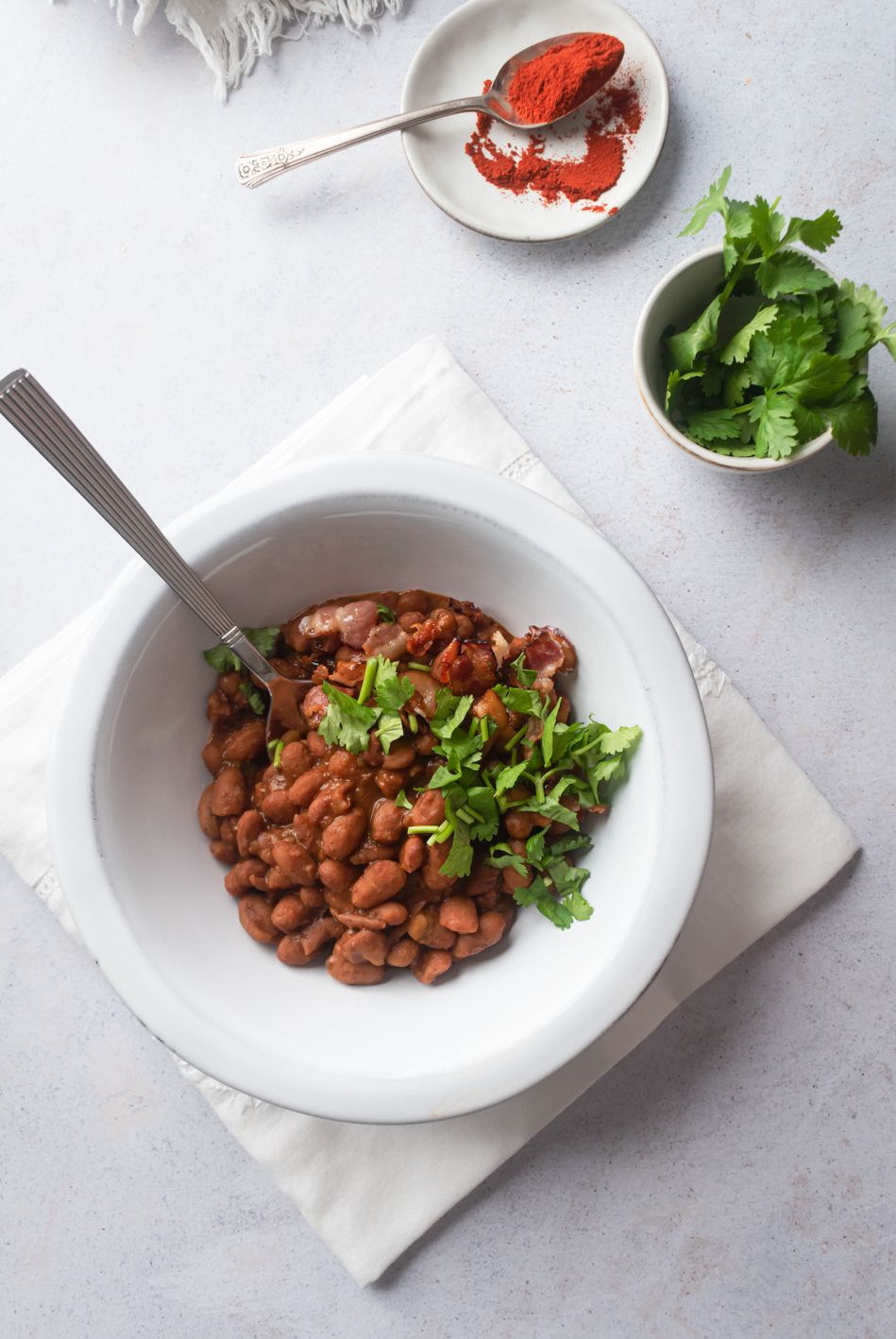 Drunken pinto beans in a white bowl with cilantro, bacon pieces, and paprika on the side.