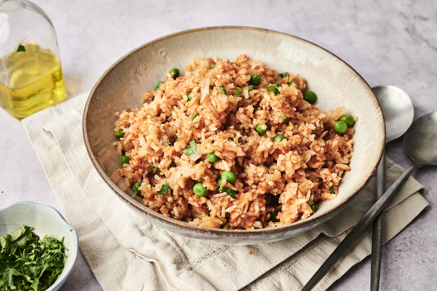 A bowl of slow cooker Mexican rice garnished with peas and chopped cilantro, served on a neutral napkin with a bottle of olive oil and a bowl of fresh herbs in the background.