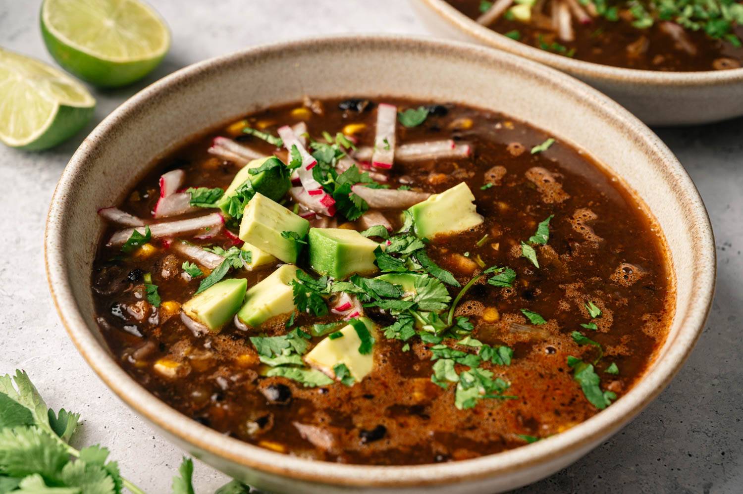 A bowl of black bean soup topped with diced avocado, radish slices, and cilantro, creating a fresh and vibrant look. Lime halves are visible nearby.