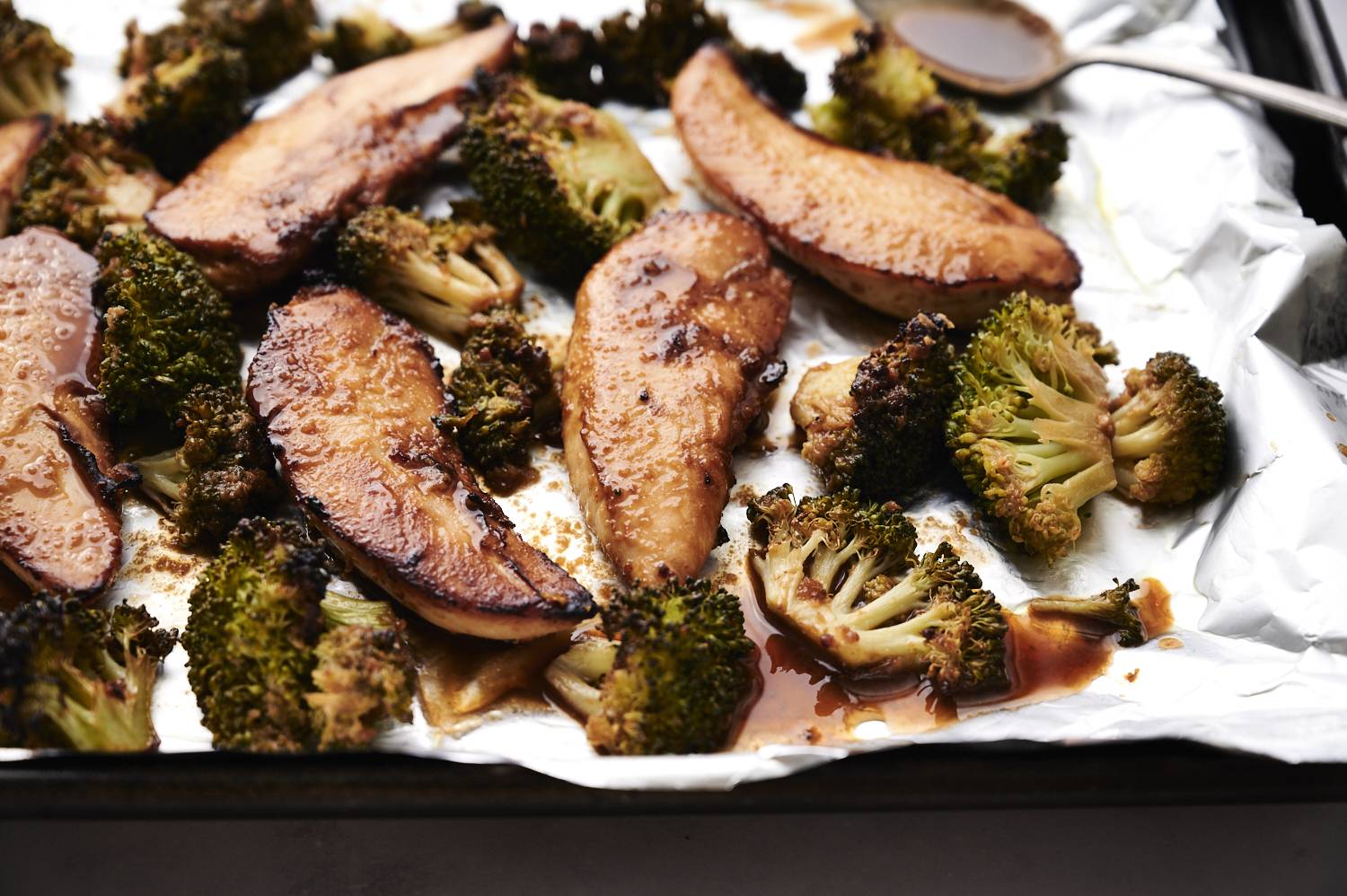 Roasted chicken slices and broccoli on a baking sheet, coated in a glossy sauce, with a silver spoon resting nearby.