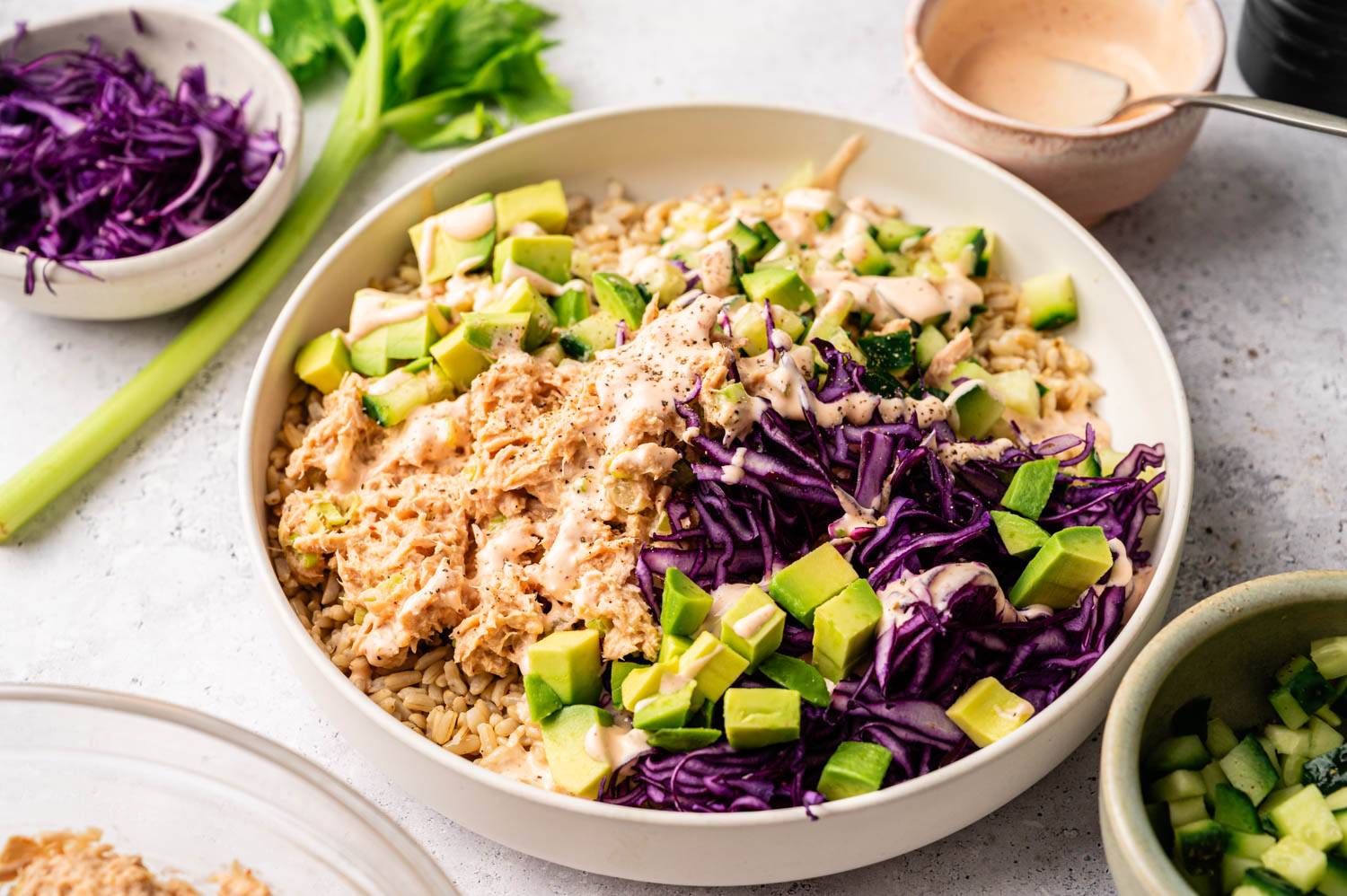 A vibrant bowl with shredded chicken, purple cabbage, avocado, and cucumber over grains, drizzled with creamy sauce, surrounded by fresh ingredients.