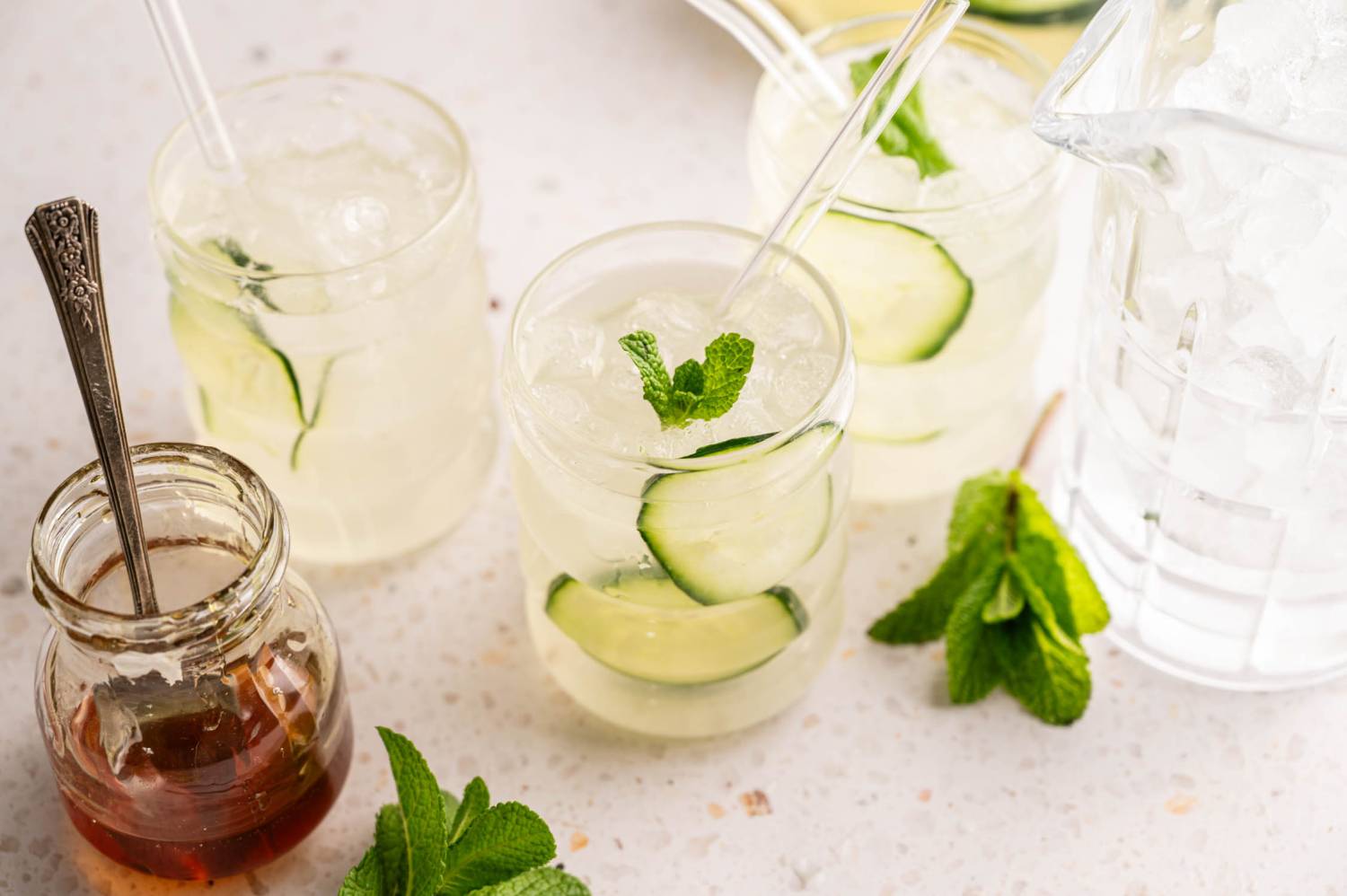 Three glasses of iced cucumber water with mint leaves and straws sit on a table. A jar of honey and fresh mint leaves are nearby on a light surface.
