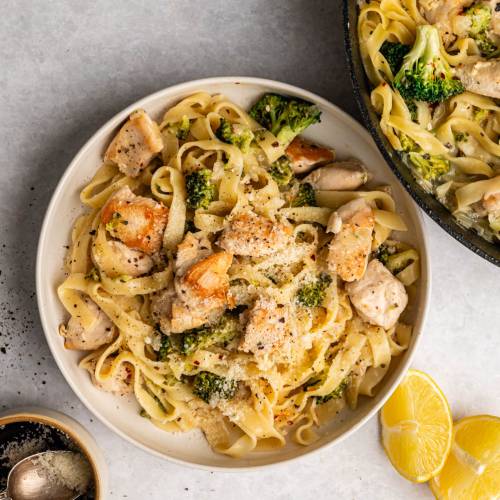 Plate of creamy chicken and broccoli pasta topped with parmesan, next to a similar skillet. Lemon wedges and pepper grinder add fresh vibes.