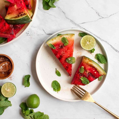 Slices of grilled watermelon on a white plate garnished with fresh basil, cut limes, and chili powder on the side.