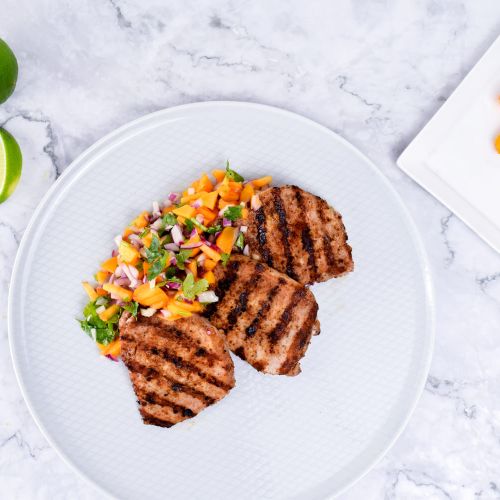 White plate with grilled pork chops accompanied with peach salsa, fresh limes and peaches on the side.