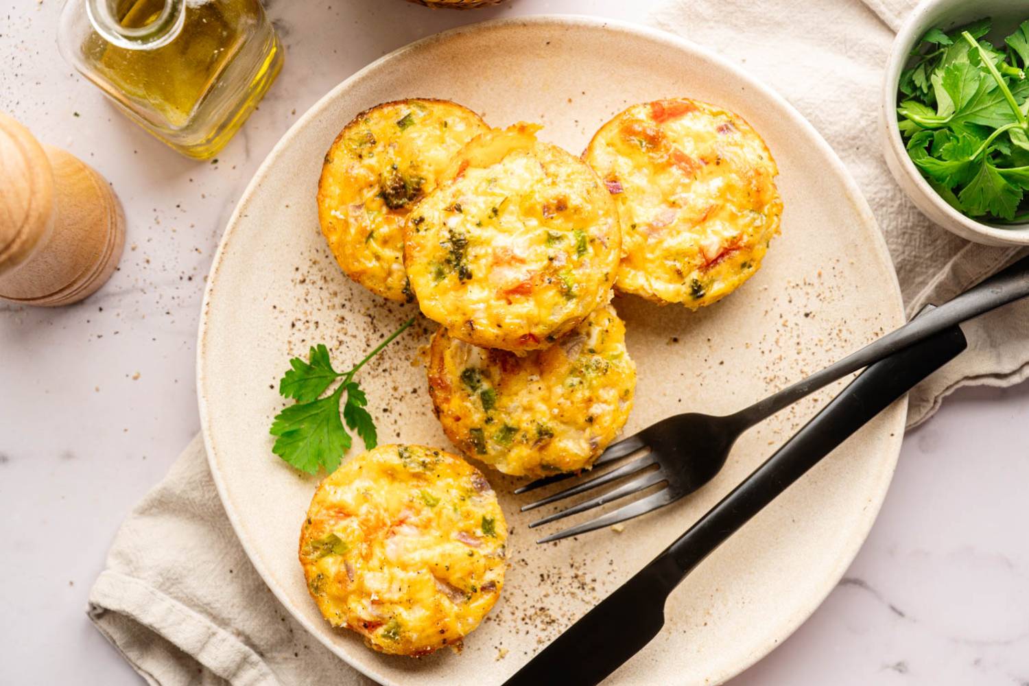 Plate of five colorful egg muffins with veggies, garnished with a parsley leaf. Accompanied by a black fork, napkin, and nearby olive oil. Inviting and fresh.