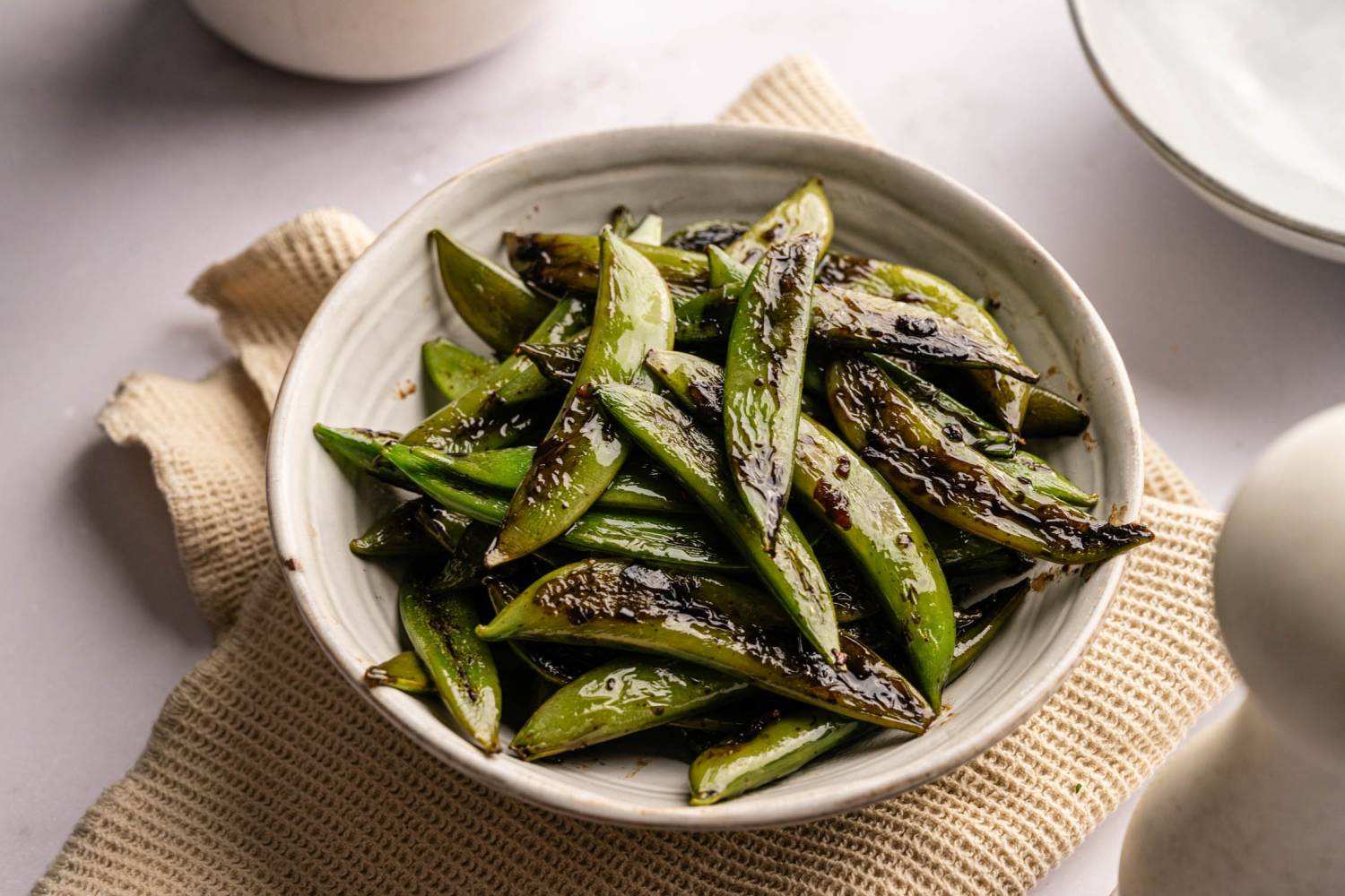 A white bowl filled with glossy, sautéed sugar snap peas on a textured beige cloth. 