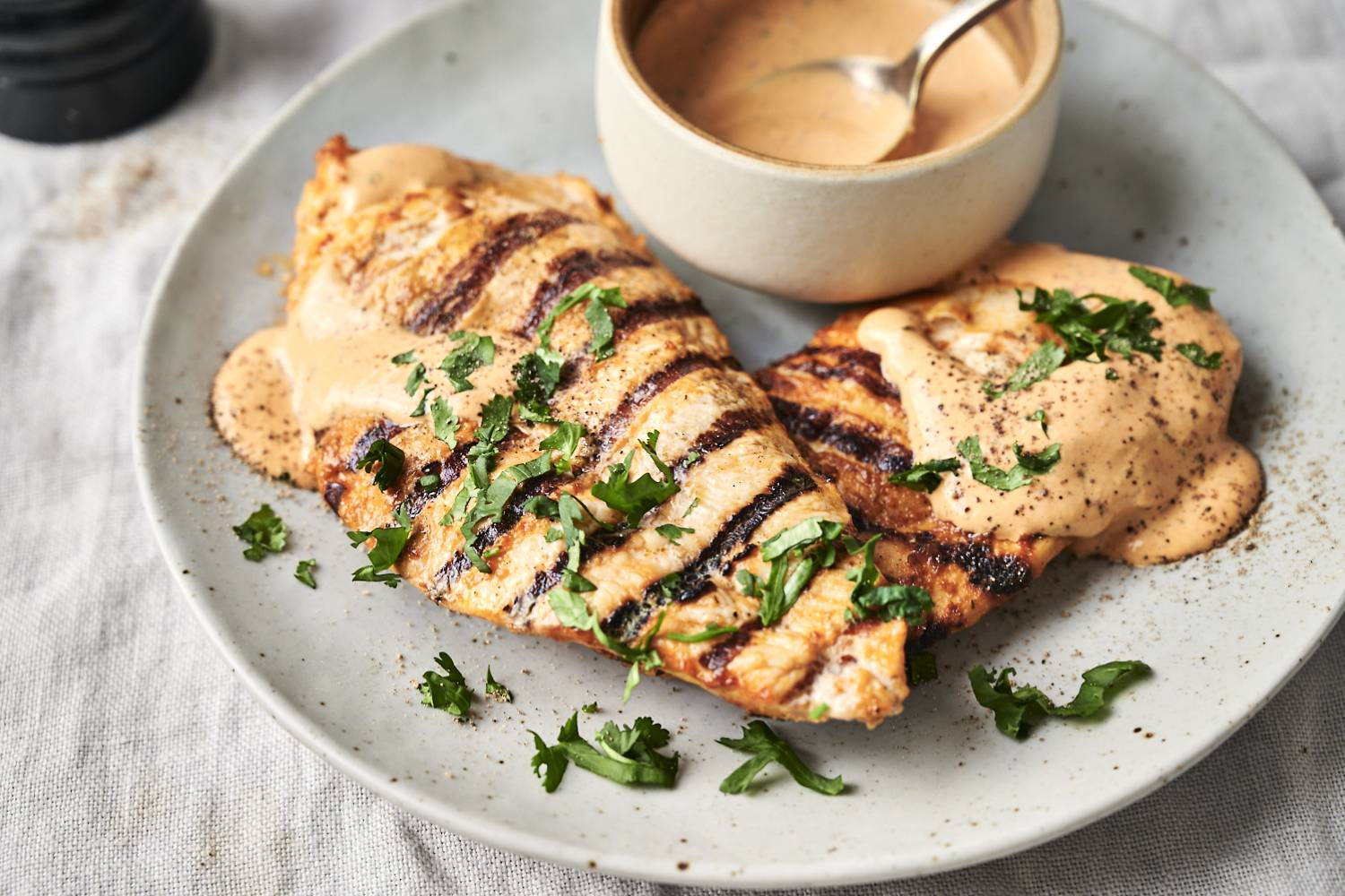 Grilled chicken breasts with char marks topped with creamy sauce and cilantro on a plate, next to a bowl of sauce with a spoon, set on a light tablecloth.