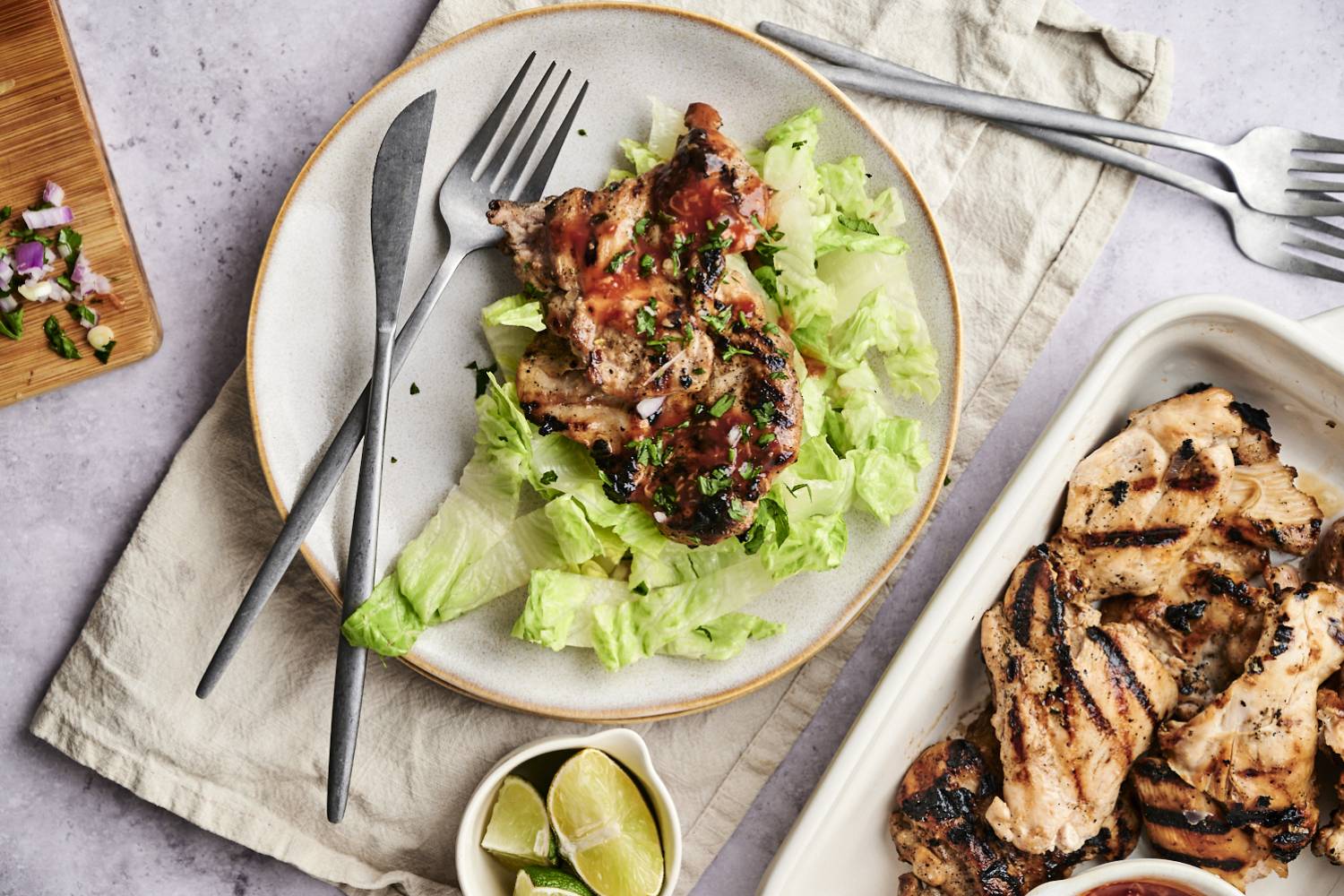 Grilled chicken topped with sauce on a bed of lettuce, served with lime wedges and garnished with cilantro, next to a platter of more chicken.
