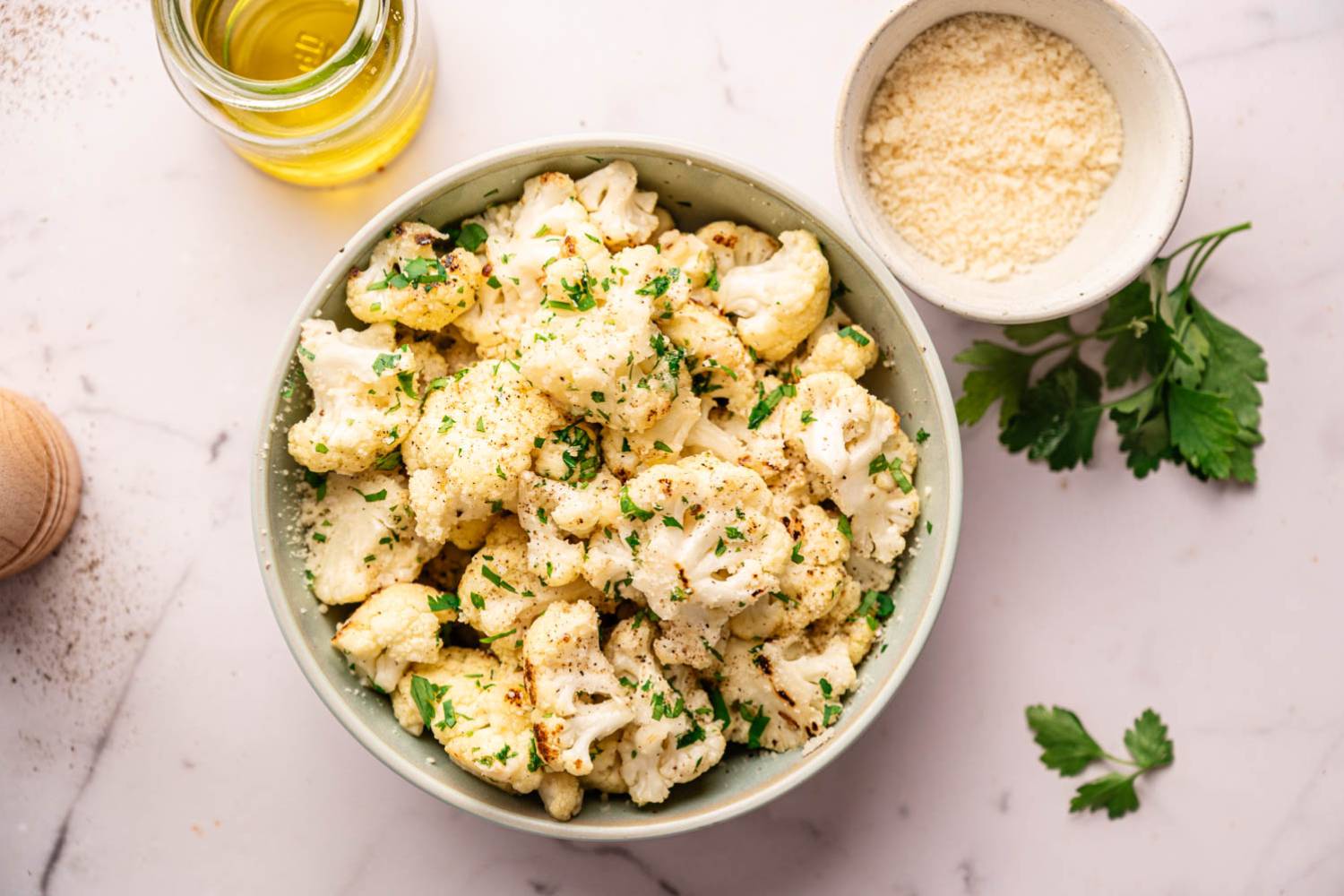 A bowl of roasted cauliflower florets garnished with chopped parsley on a marble surface. Nearby are a bowl of grated cheese and a jar of olive oil.