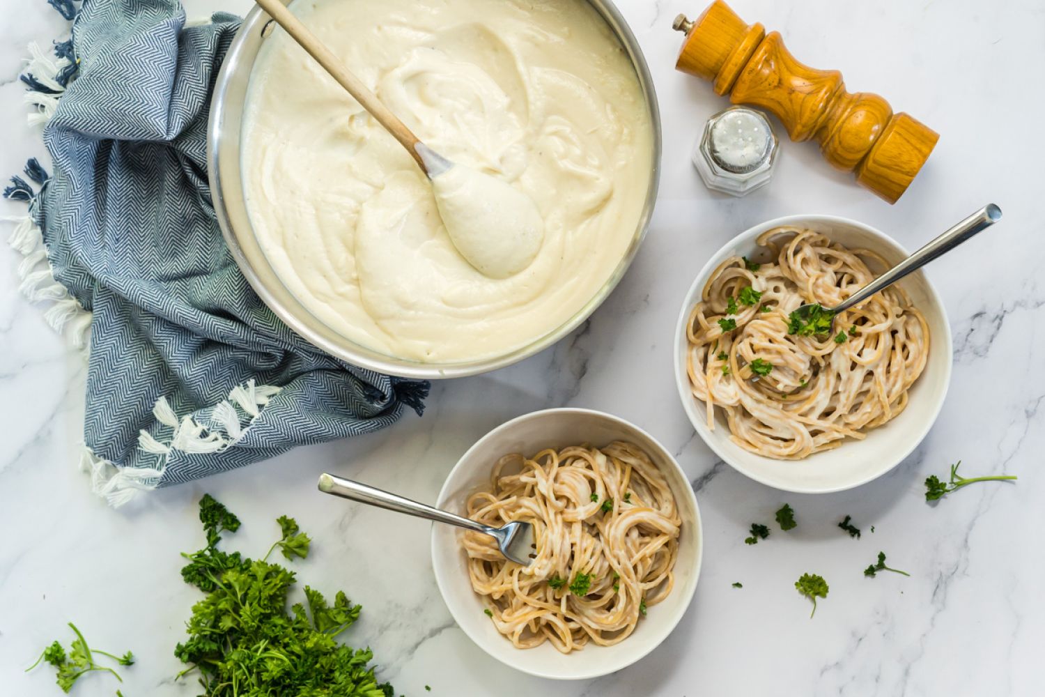 Skinny Alfredo sauce in a large pan with a wooden spoon and served in two bowls with spaghetti and parsley.