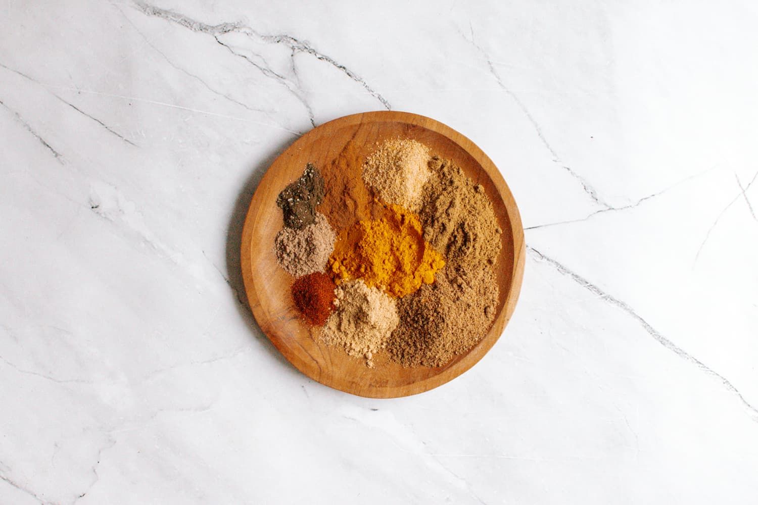 Homemade curry powder on a wooden plate with a variety of ground spices including cumin, coriander, cardamom, ginger, cinnamon, and cayenne.