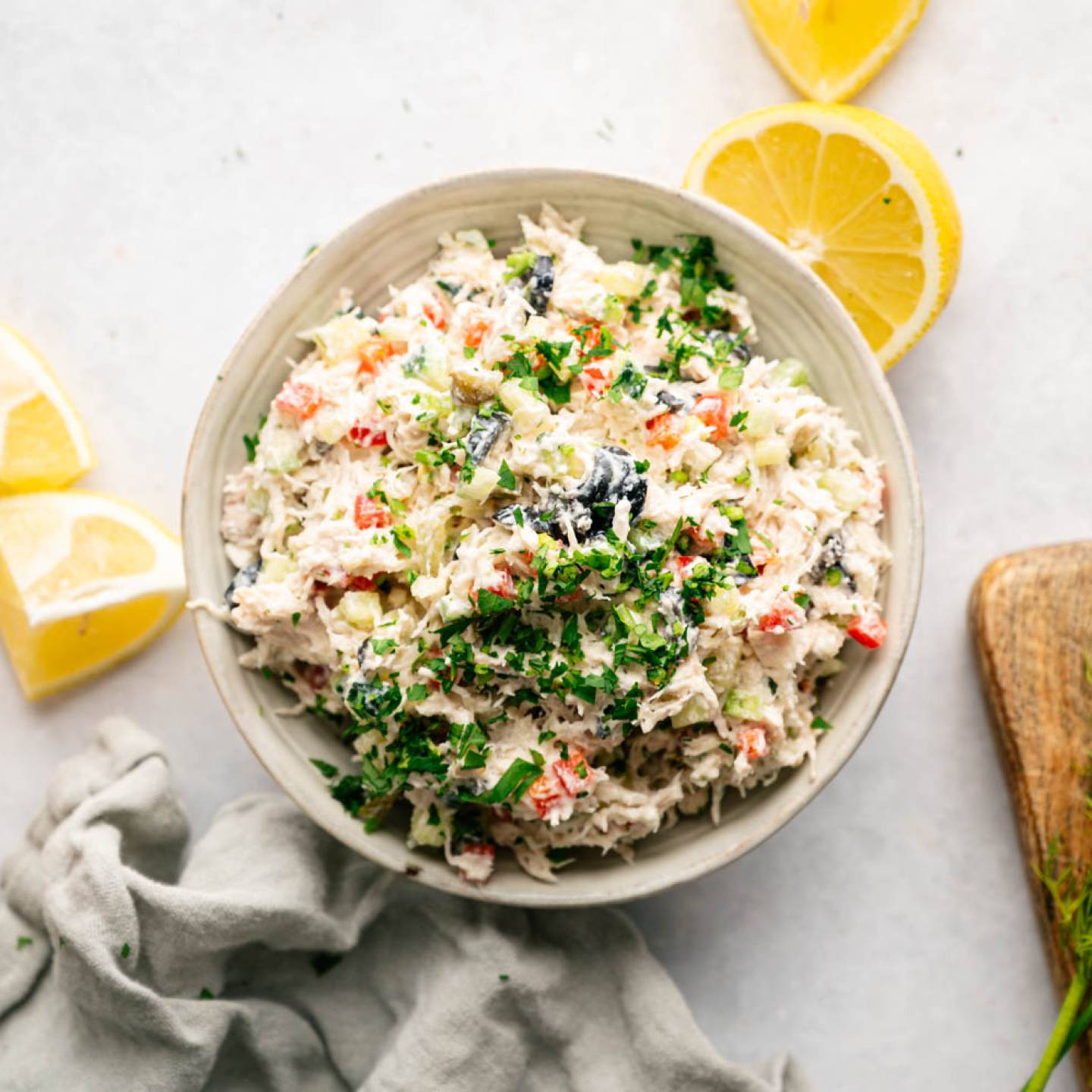 A bowl of colorful crab salad garnished with fresh herbs, surrounded by lemon wedges, dill sprigs, a wooden board, and a gray cloth on a light surface.