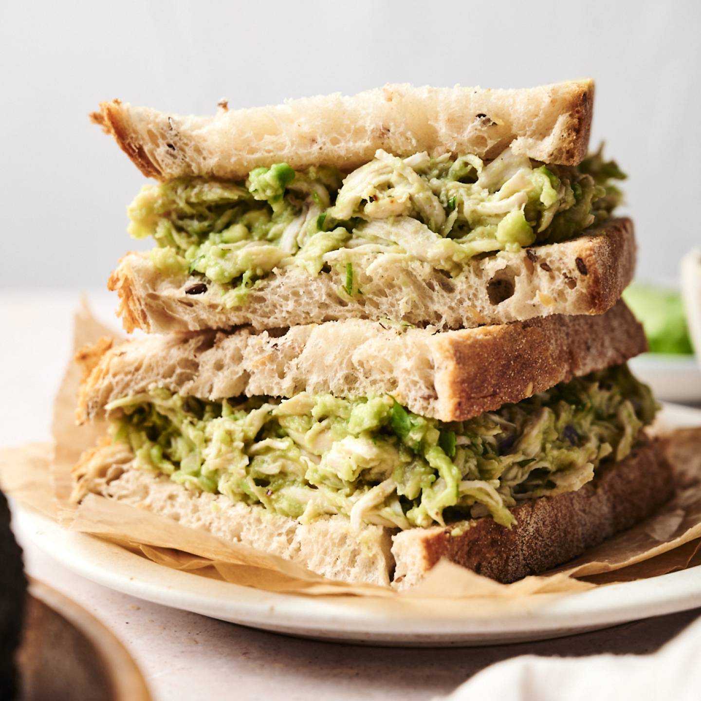 Close-up of a stacked sandwich made with thick slices of rustic bread and filled generously with creamy avocado chicken salad. The sandwich is served on a parchment-lined plate, ready to enjoy.