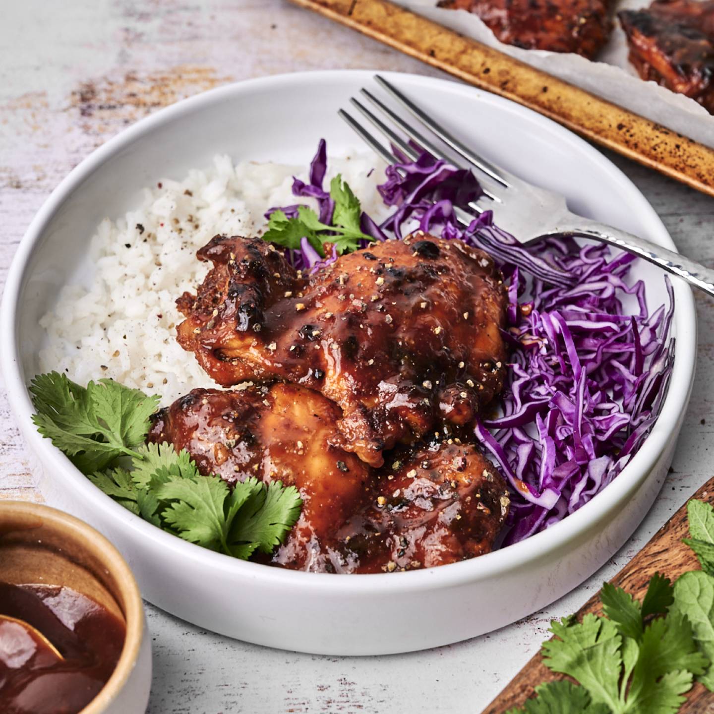 A white bowl filled with baked BBQ chicken thighs glazed in a rich sauce, served alongside fluffy white rice, shredded purple cabbage, and fresh cilantro, with a fork resting on the side.