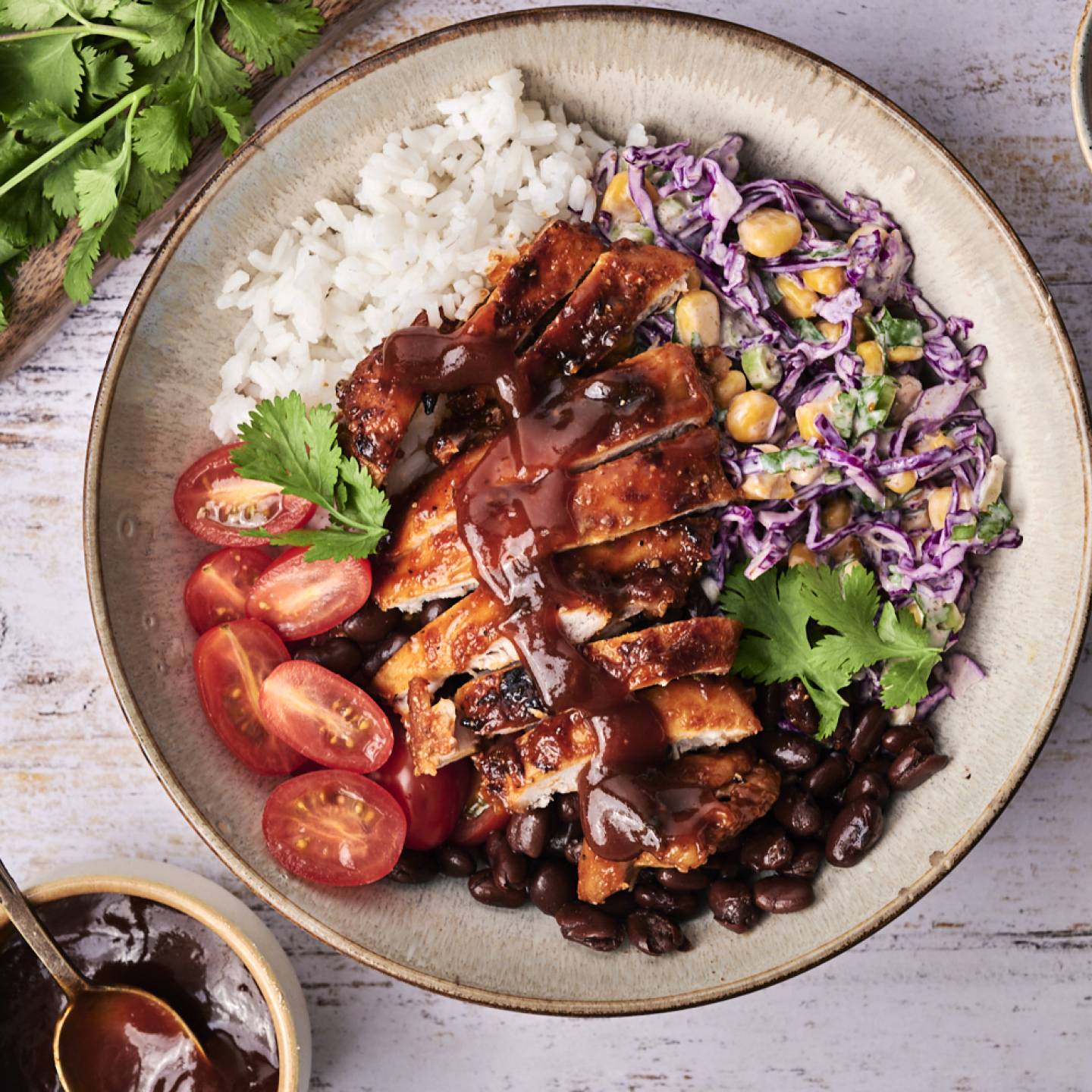 A colorful bowl featuring grilled chicken, rice, black beans, cherry tomatoes, slaw, and herbs, drizzled with barbecue sauce.