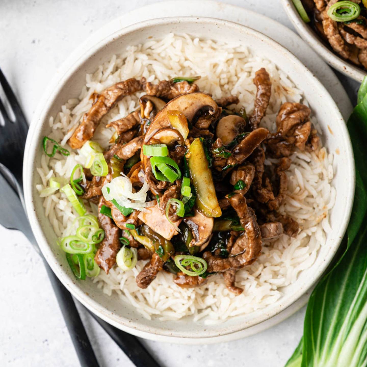 A bowl of rice topped with stir-fried beef, mushrooms, zucchini, and green onions. Nearby are black cutlery and fresh bok choy leaves, suggesting an Asian-inspired meal.