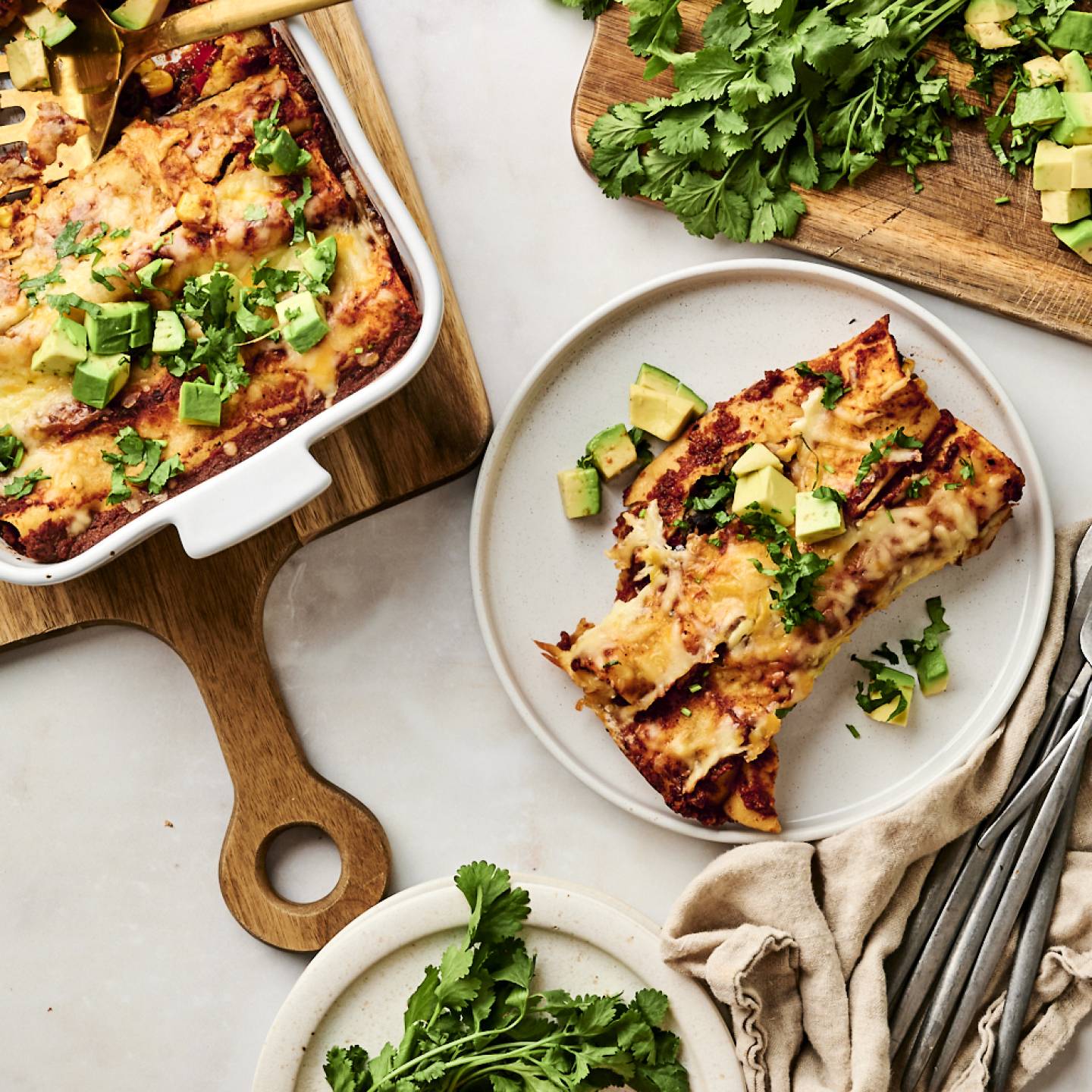 Black Bean Enchiladas Cheesy black bean enchiladas topped with diced avocado and fresh cilantro, served on a white plate with a side of extra herbs on a wooden cutting board.