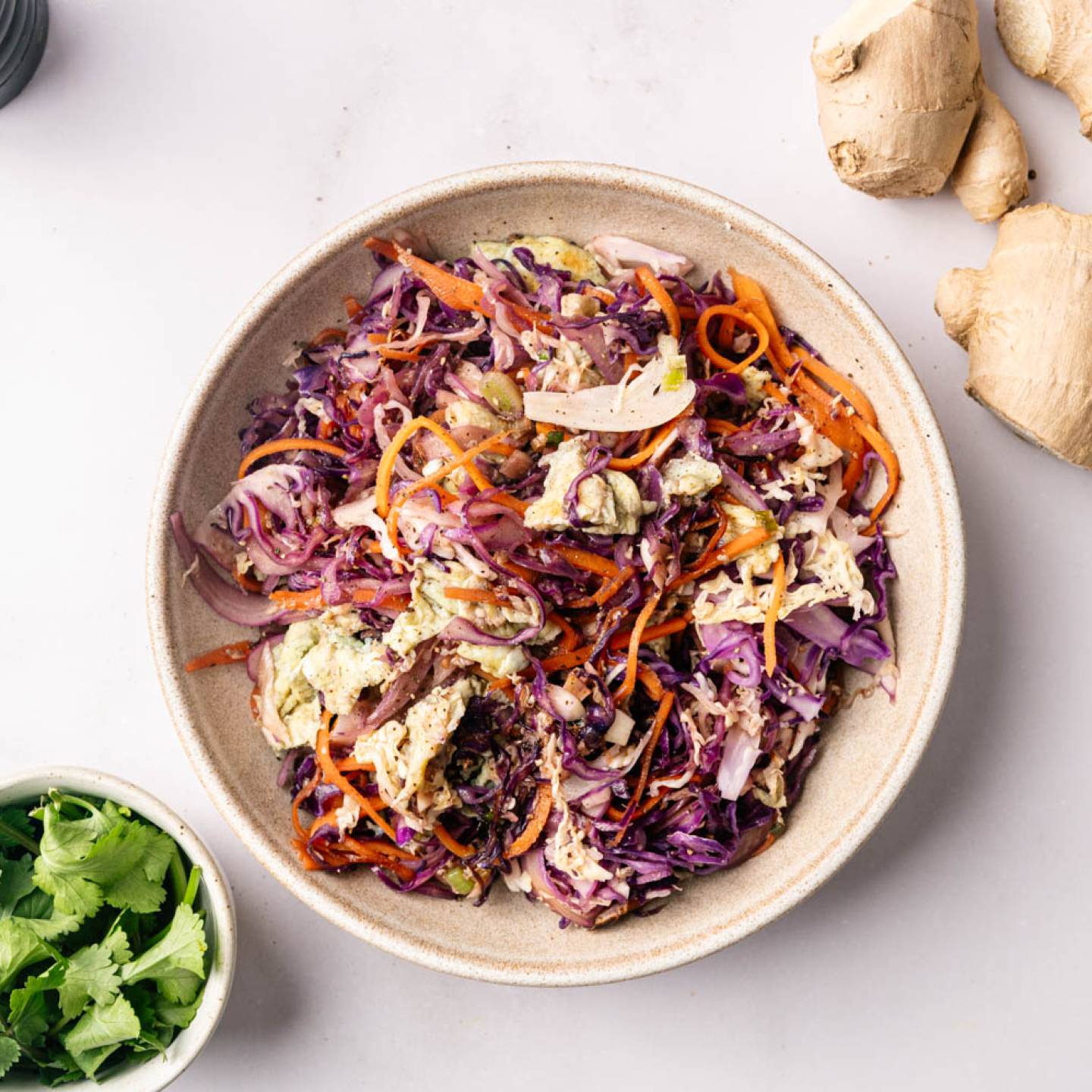 A colorful salad with shredded purple cabbage, carrots, and dressing in a beige bowl on a white surface. Fresh cilantro and ginger root surround it, suggesting fresh ingredients.