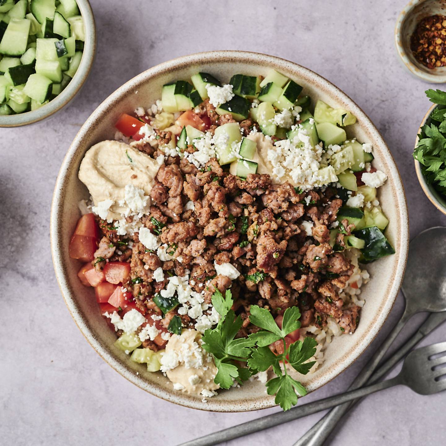 Bowl of hummus topped with seasoned ground meat, cucumber, tomatoes, feta, and fresh parsley, surrounded by ingredient bowls and utensils.