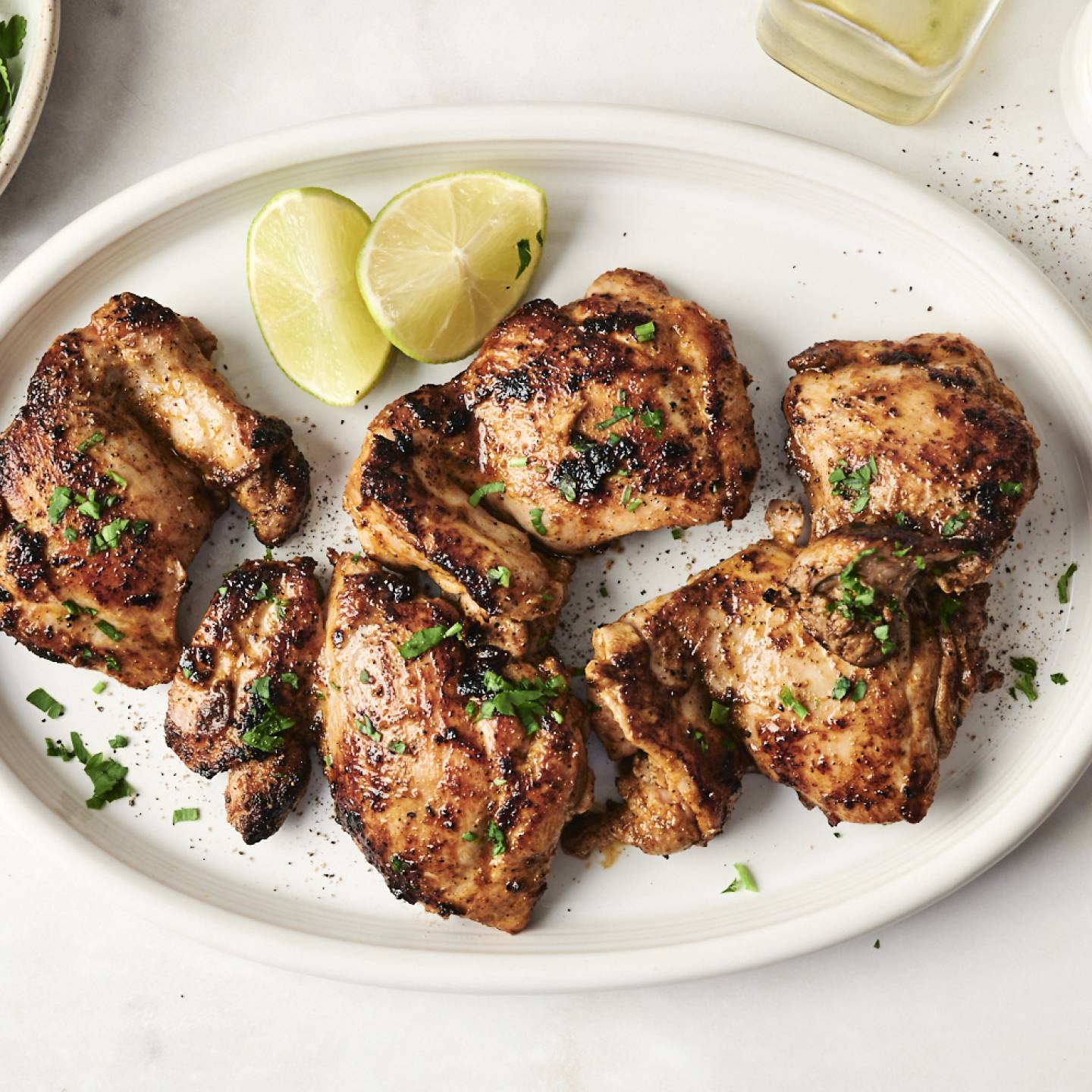 Juicy chipotle chicken thighs served on a white platter, garnished with fresh cilantro and lime wedges, surrounded by bowls of salt, herbs, and olive oil for a vibrant presentation.