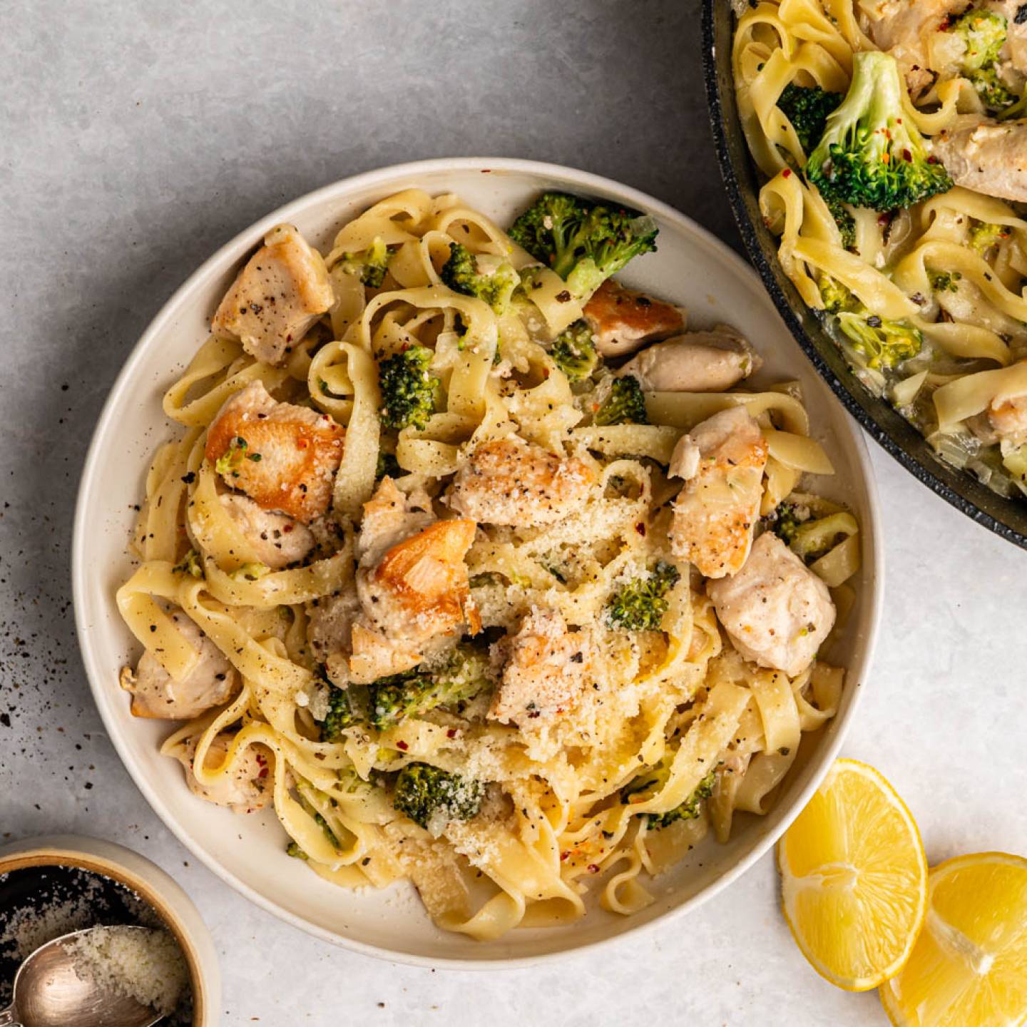Plate of creamy chicken and broccoli pasta topped with parmesan, next to a similar skillet. Lemon wedges and pepper grinder add fresh vibes.