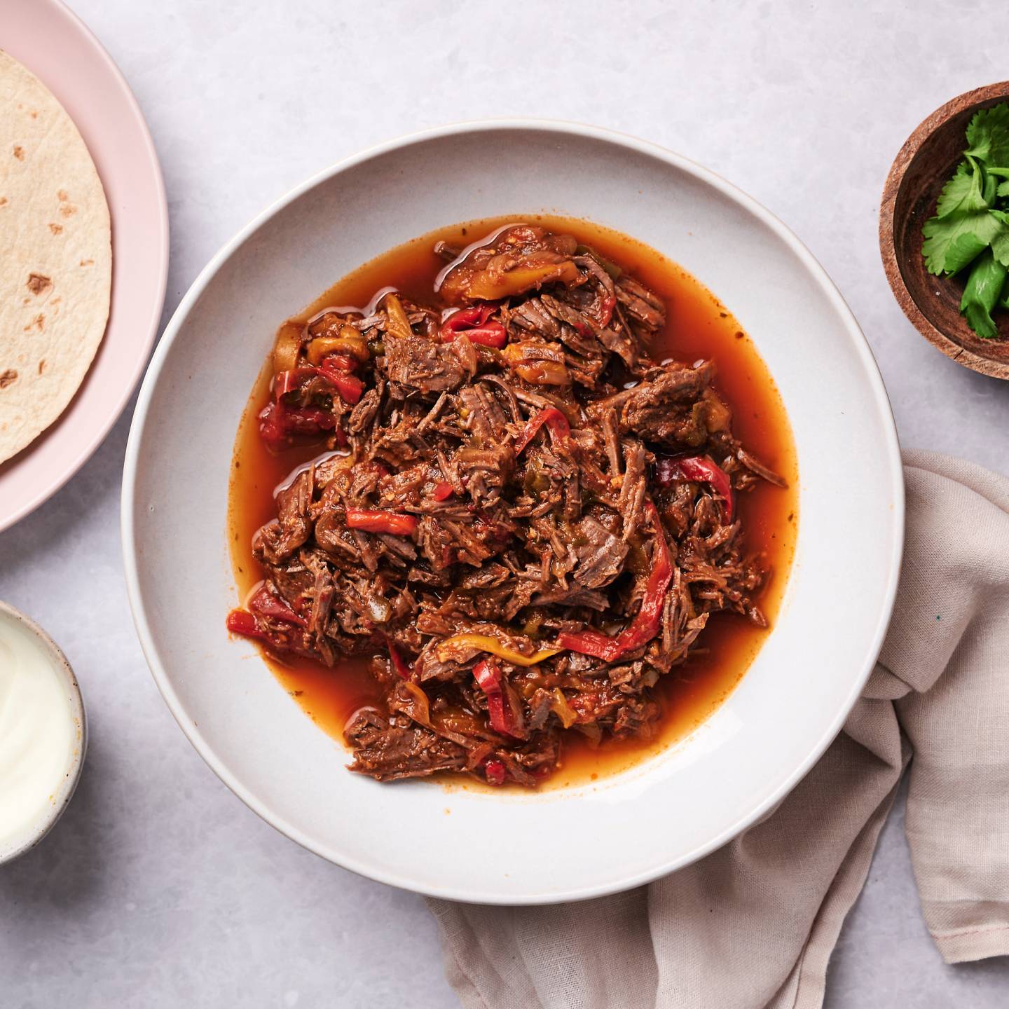Crockpot beef fajitas with bell peppers, onions, and fajita sauce in a bowl with tortillas, cilantro, and sour cream on the side.
