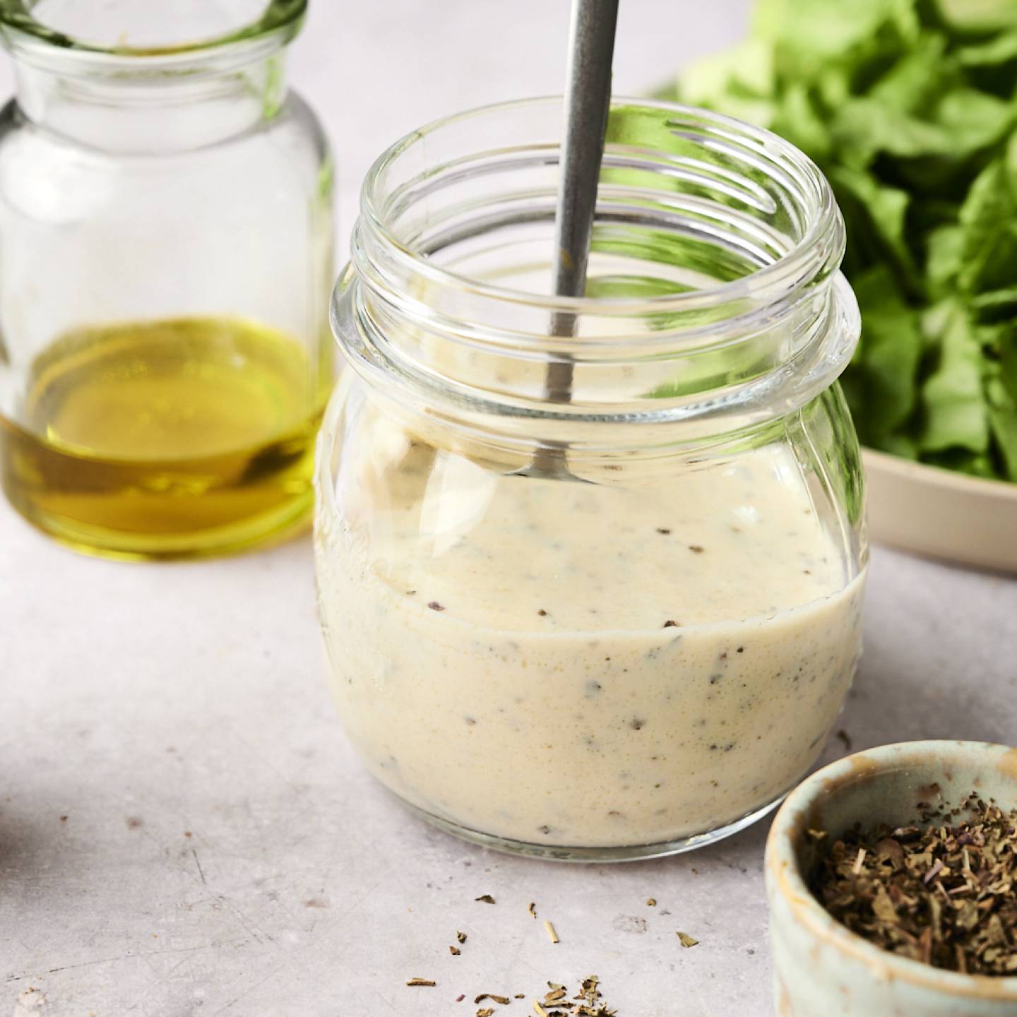 Jar of creamy Greek salad dressing with herbs, olive oil, and fresh lettuce in the background.