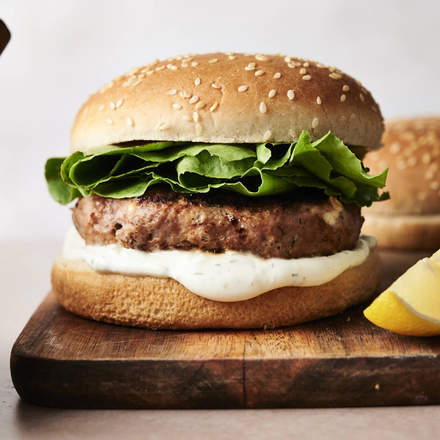 Greek turkey burger on a wooden board with crisp lettuce and creamy tzatziki sauce inside a sesame seed bun, garnished with a lemon wedge on the side.