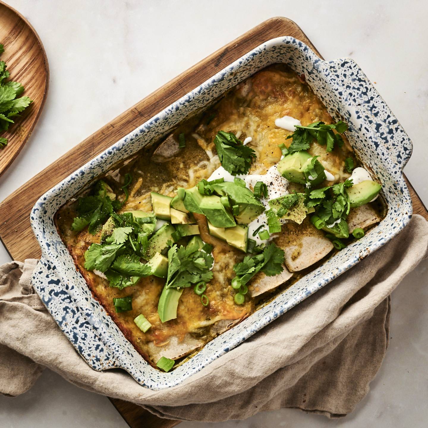 A baking dish filled with green chicken enchiladas, topped with melted cheese, avocado, cilantro, sour cream, and green onions, placed on a wooden cutting board with fresh cilantro and jalapeños on the side.