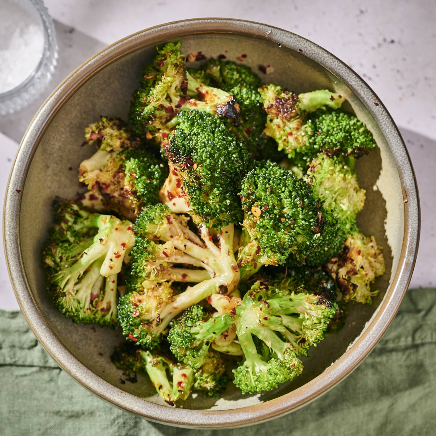 A bowl of roasted broccoli with crispy edges, sprinkled with red pepper flakes, sits on a light surface beside a salt dish.