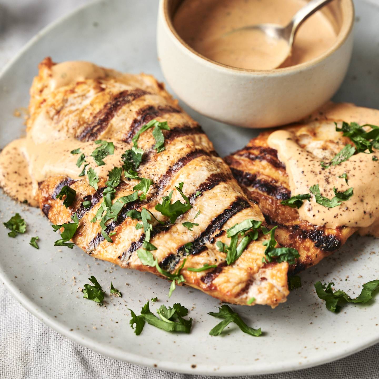 Grilled chicken breasts with char marks topped with creamy sauce and cilantro on a plate, next to a bowl of sauce with a spoon, set on a light tablecloth.