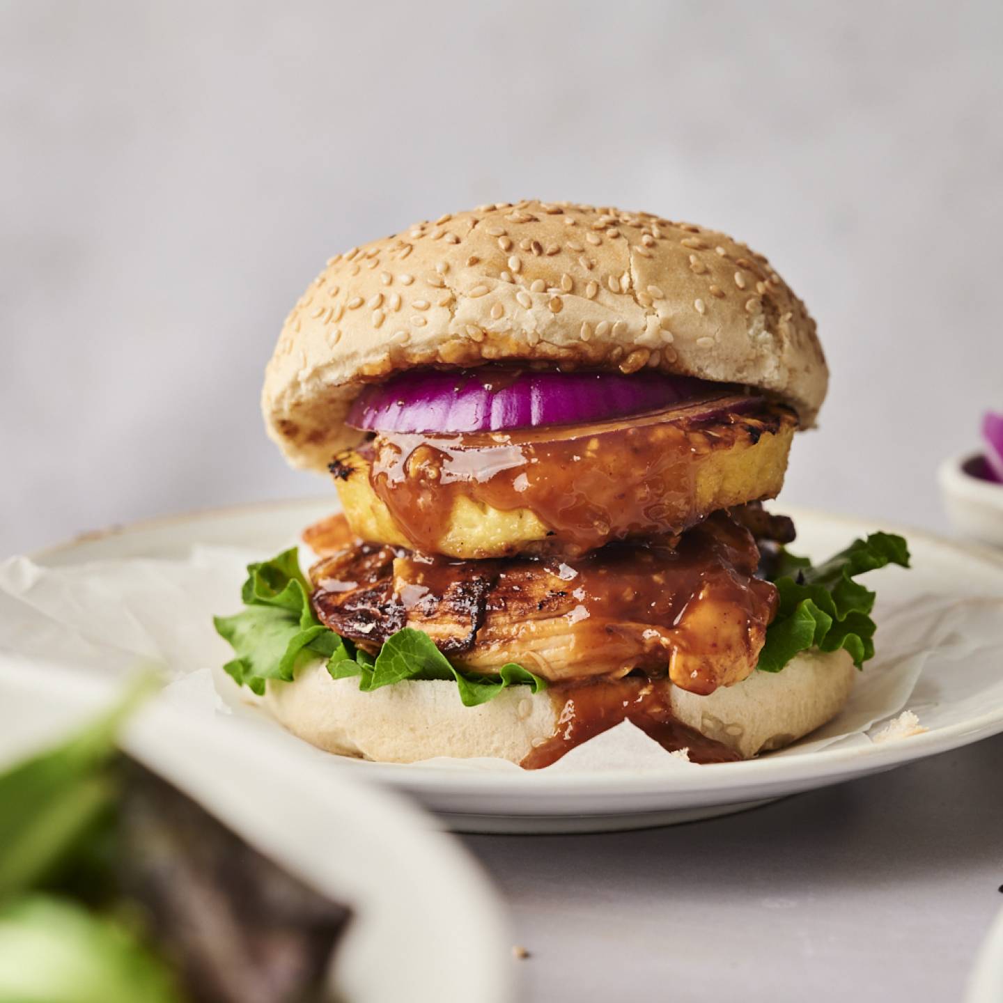 A delicious sesame seed burger with grilled chicken, lettuce, a slice of pineapple, red onion, and a savory sauce on a white plate.