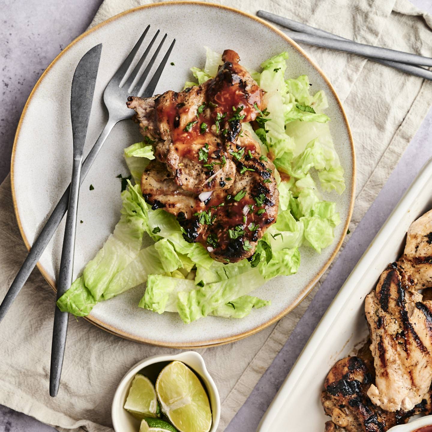 Grilled chicken topped with sauce on a bed of lettuce, served with lime wedges and garnished with cilantro, next to a platter of more chicken.