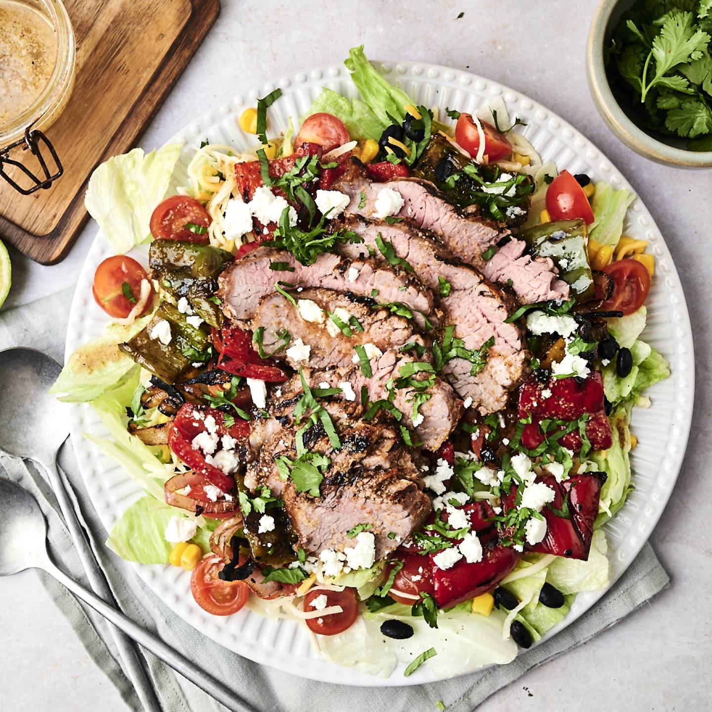 A vibrant salad topped with sliced grilled beef, assorted veggies, feta cheese, and cilantro, accompanied by lime halves and dressing.
