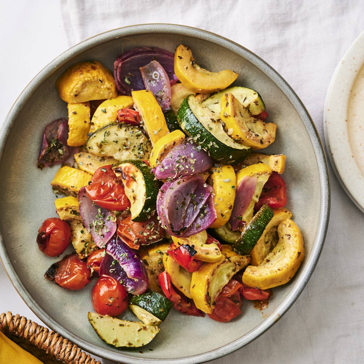 A bowl of colorful roasted vegetables, including zucchini, yellow squash, and cherry tomatoes, garnished with herbs. Lemon slices nearby.