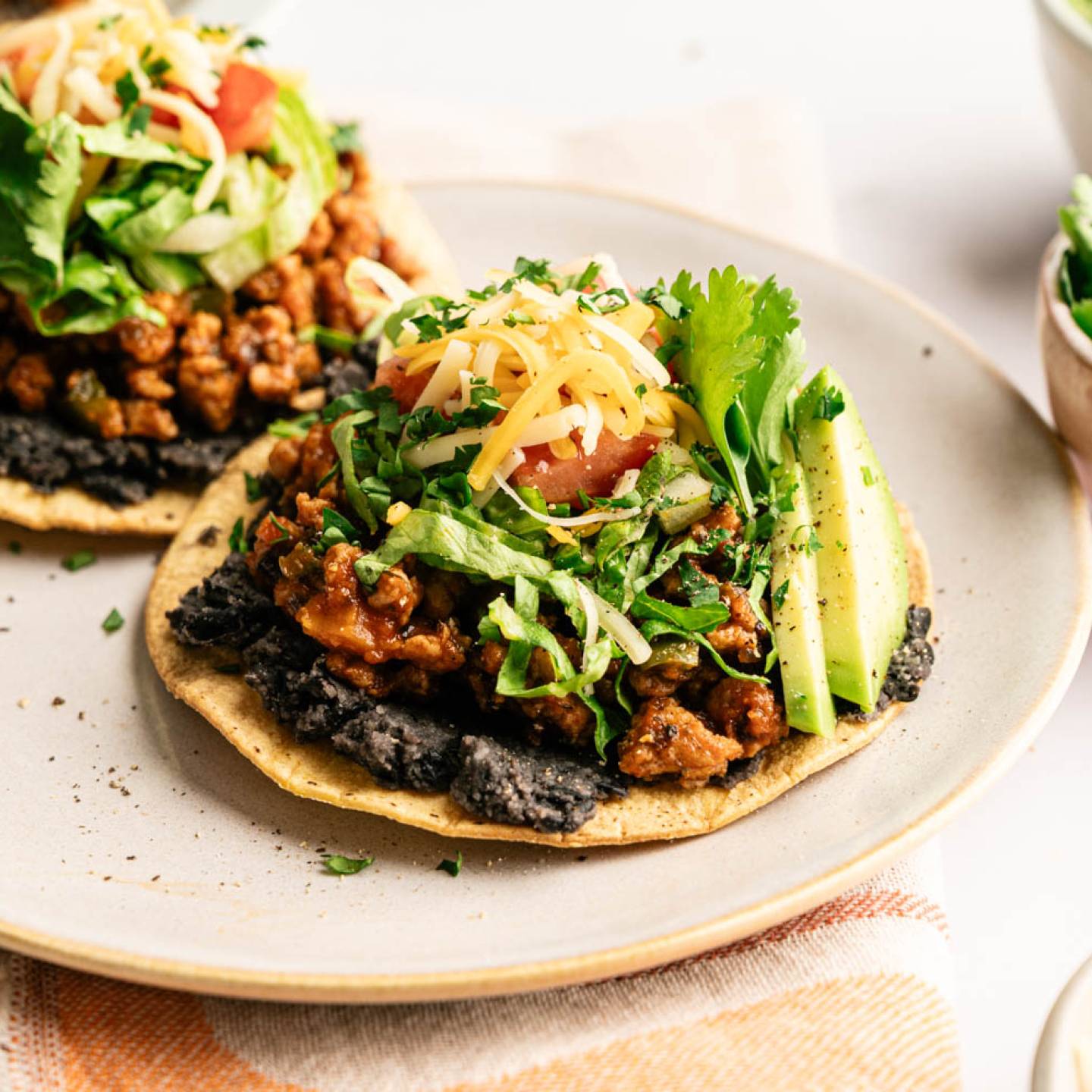 Loaded ground turkey tostadas topped with black beans, seasoned turkey, shredded lettuce, diced tomatoes, shredded cheese, fresh cilantro, and avocado slices, served on a plate with extra toppings nearby.