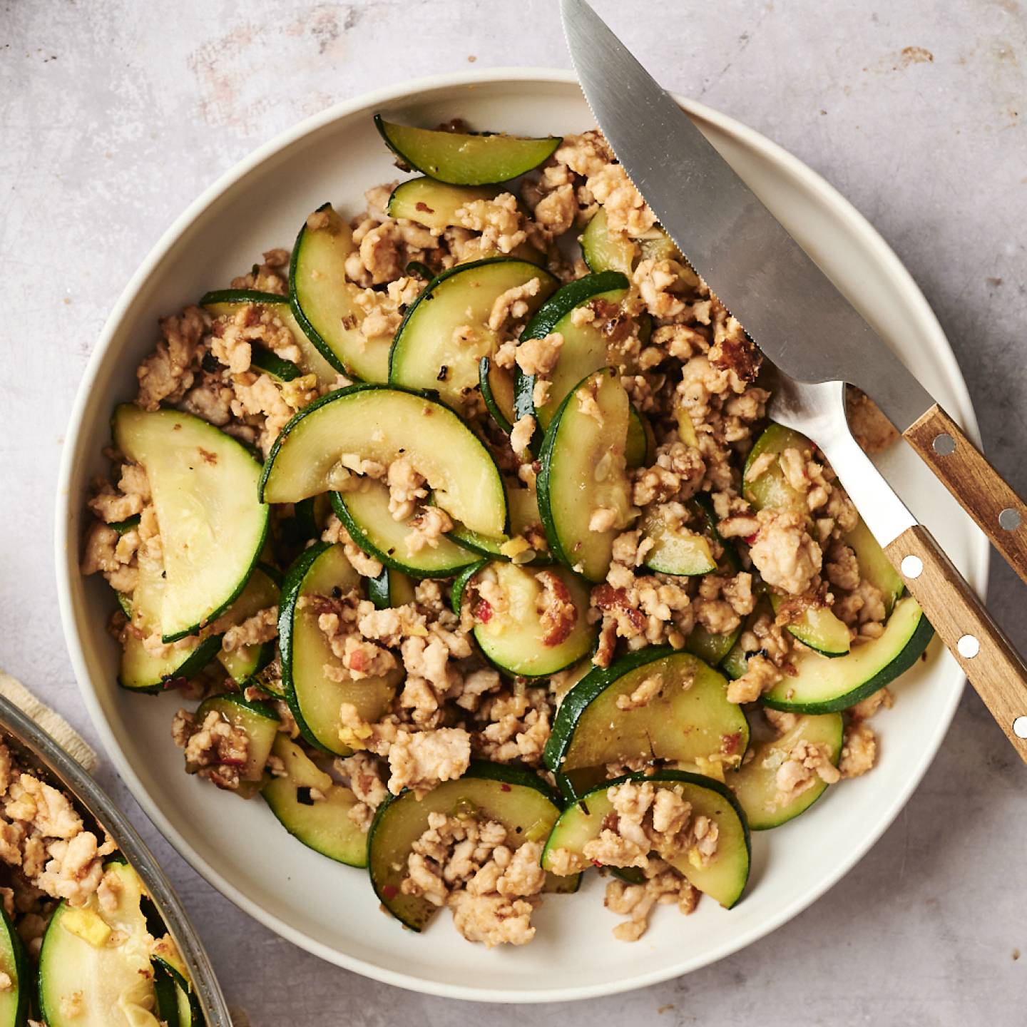 Bowl of ground turkey zucchini stir fry with tender zucchini slices and browned turkey, served with a knife and fork on the side.