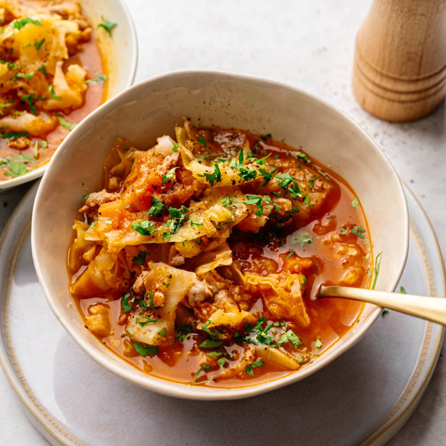 A bowl of hearty cabbage soup with ground meat, garnished with fresh parsley, sits on a light gray surface. A second bowl and pepper mill in the background.