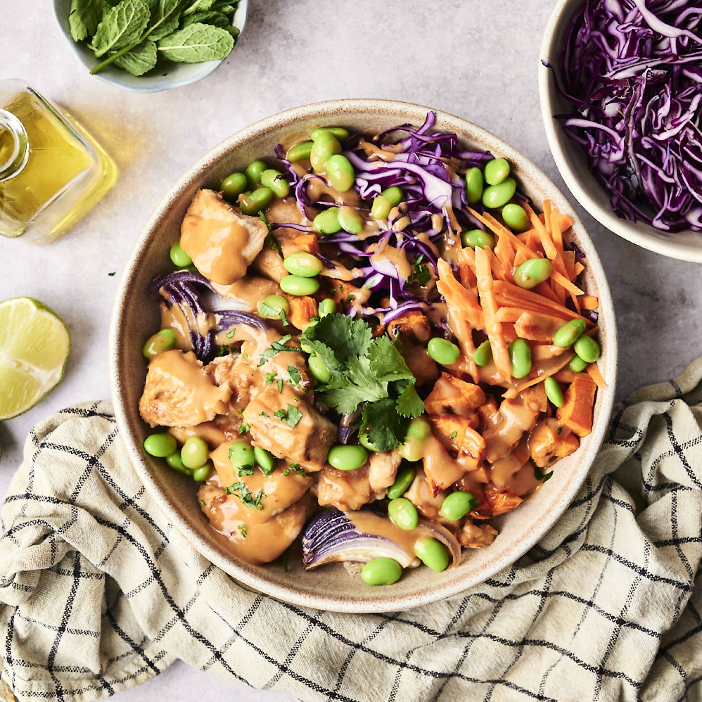 A vibrant bowl of chicken, edamame, purple cabbage, and carrots drizzled with sauce, surrounded by lime, mint, and olive oil.