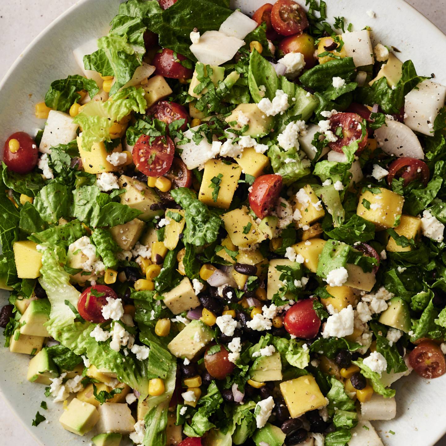 A vibrant Mexican chopped salad filled with diced mango, avocado, cherry tomatoes, black beans, corn, jicama, romaine lettuce, and crumbled cheese, garnished with fresh cilantro on a white plate.