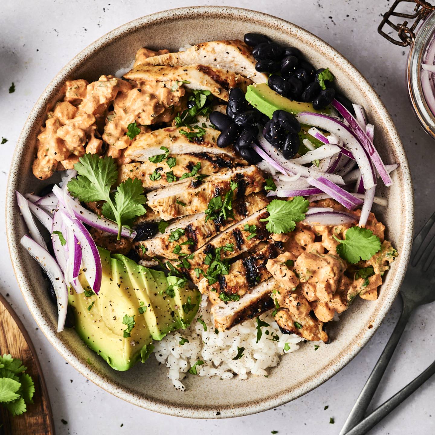 A bowl filled with grilled chicken, rice, black beans, avocado, red onion, and creamy sauce, garnished with fresh cilantro.